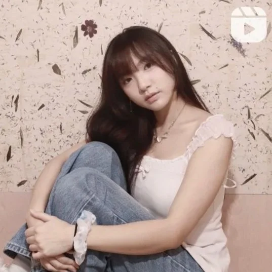 Young woman with long dark hair sitting against a textured wall with floral decorations, wearing a white top and jeans.