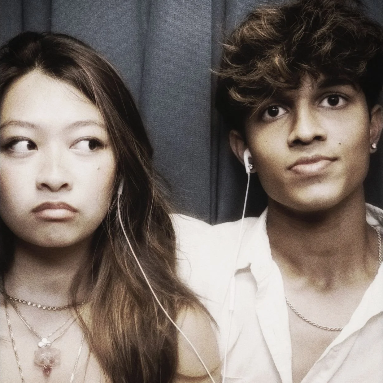A woman and a man sitting side by side, both wearing earbuds, in an indoor setting. The woman has long brown hair, and the man has curly brown hair. They appear to be sitting close together.