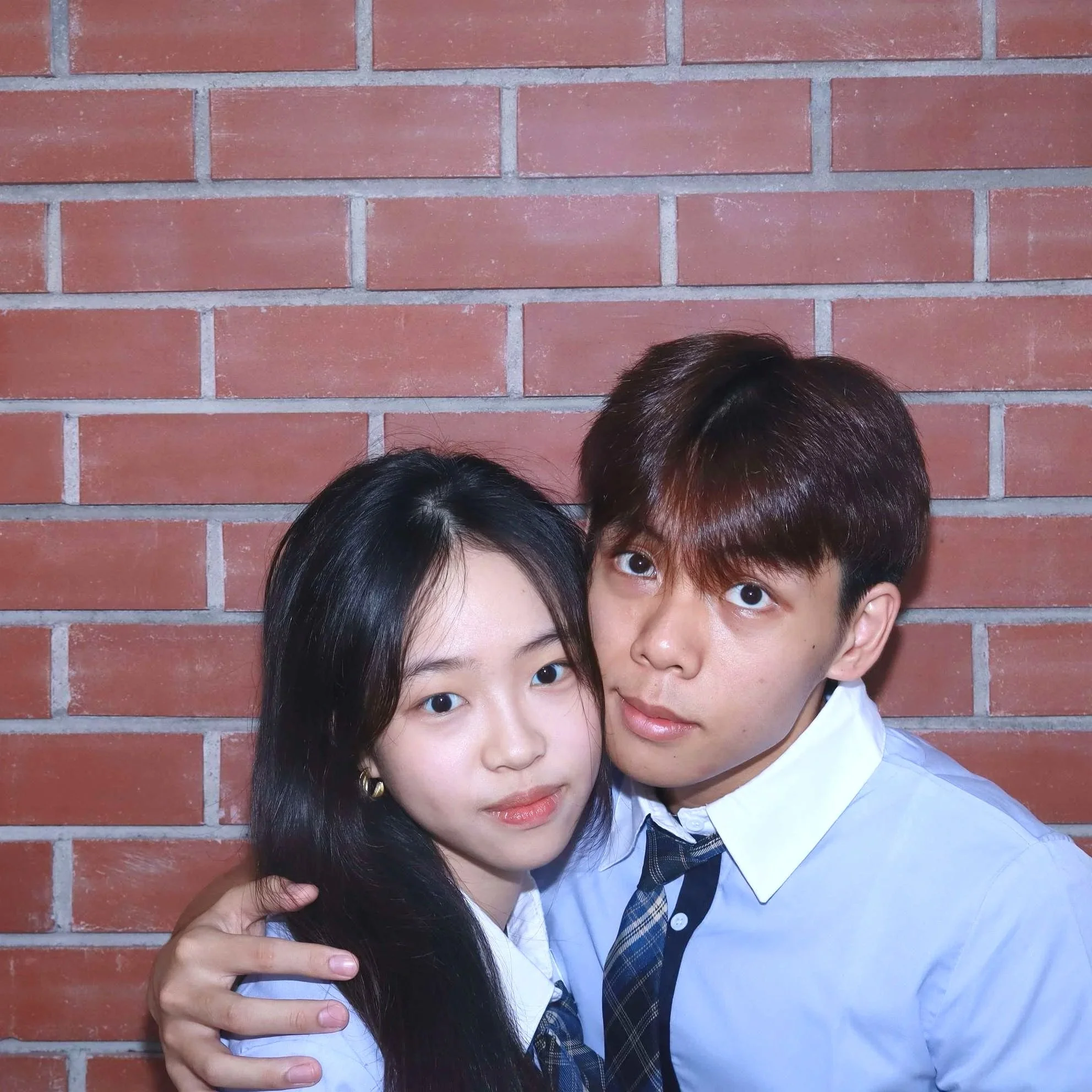 A photo of a couple in school uniforms posing against a red brick wall in Photomanner The Brick House, with one person's arm around the other's shoulder.