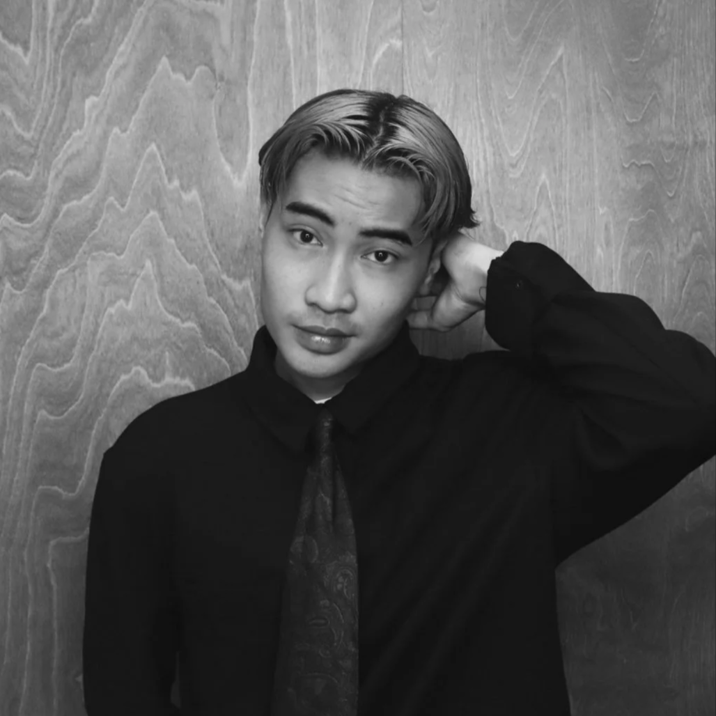 A young man with styled hair, wearing a dark shirt and tie, posing with one hand behind his head against a wooden background.