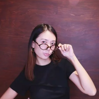 Young woman with shoulder-length hair, wearing glasses and a black shirt, posing with her glasses slightly lowered, against a wooden background.