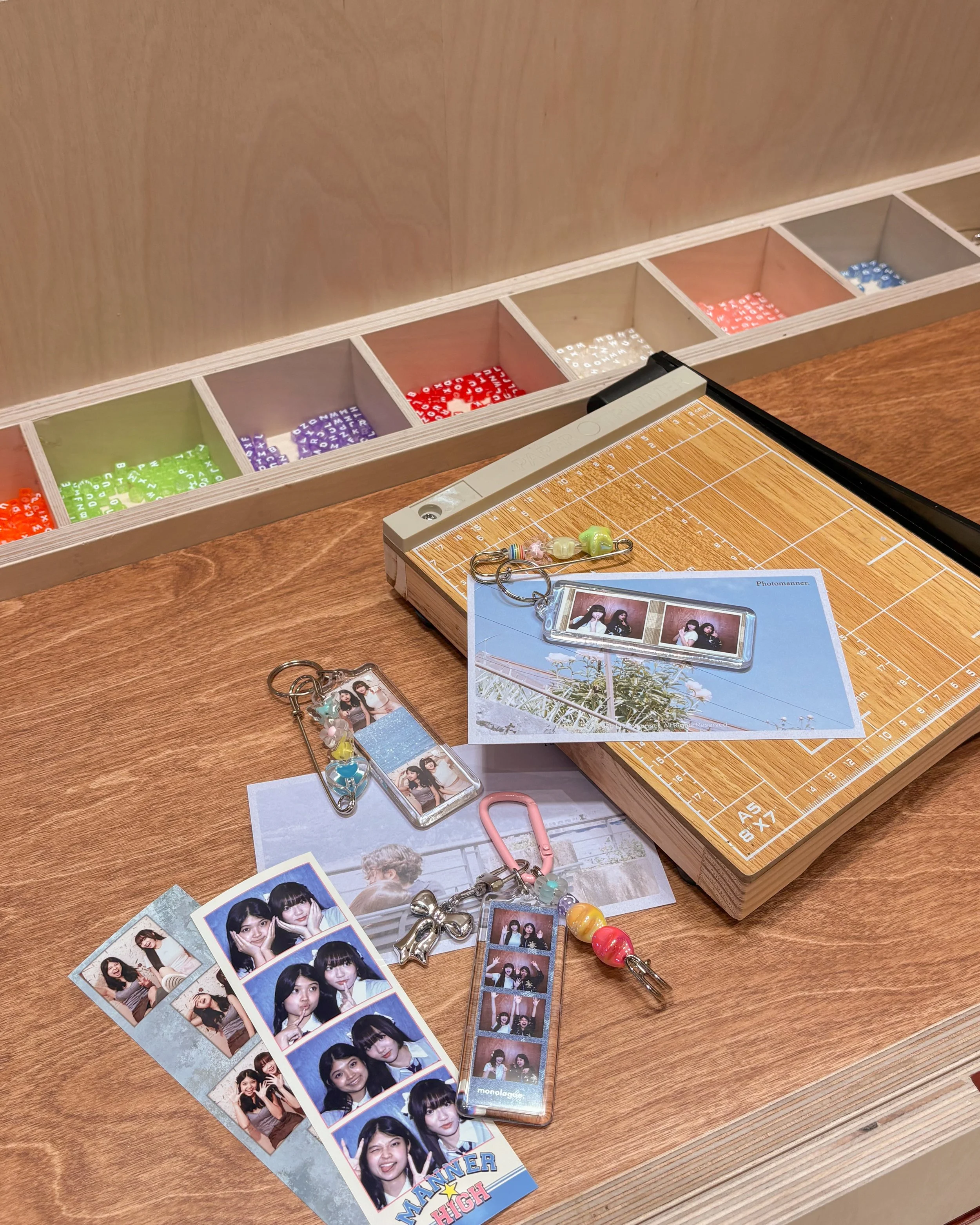Photo of photo booth pictures of young women making various facial expressions, a photo frame with a picture of two women, and decorative charms on a wooden table, with a photo strip holder and unopened photo film in the background.