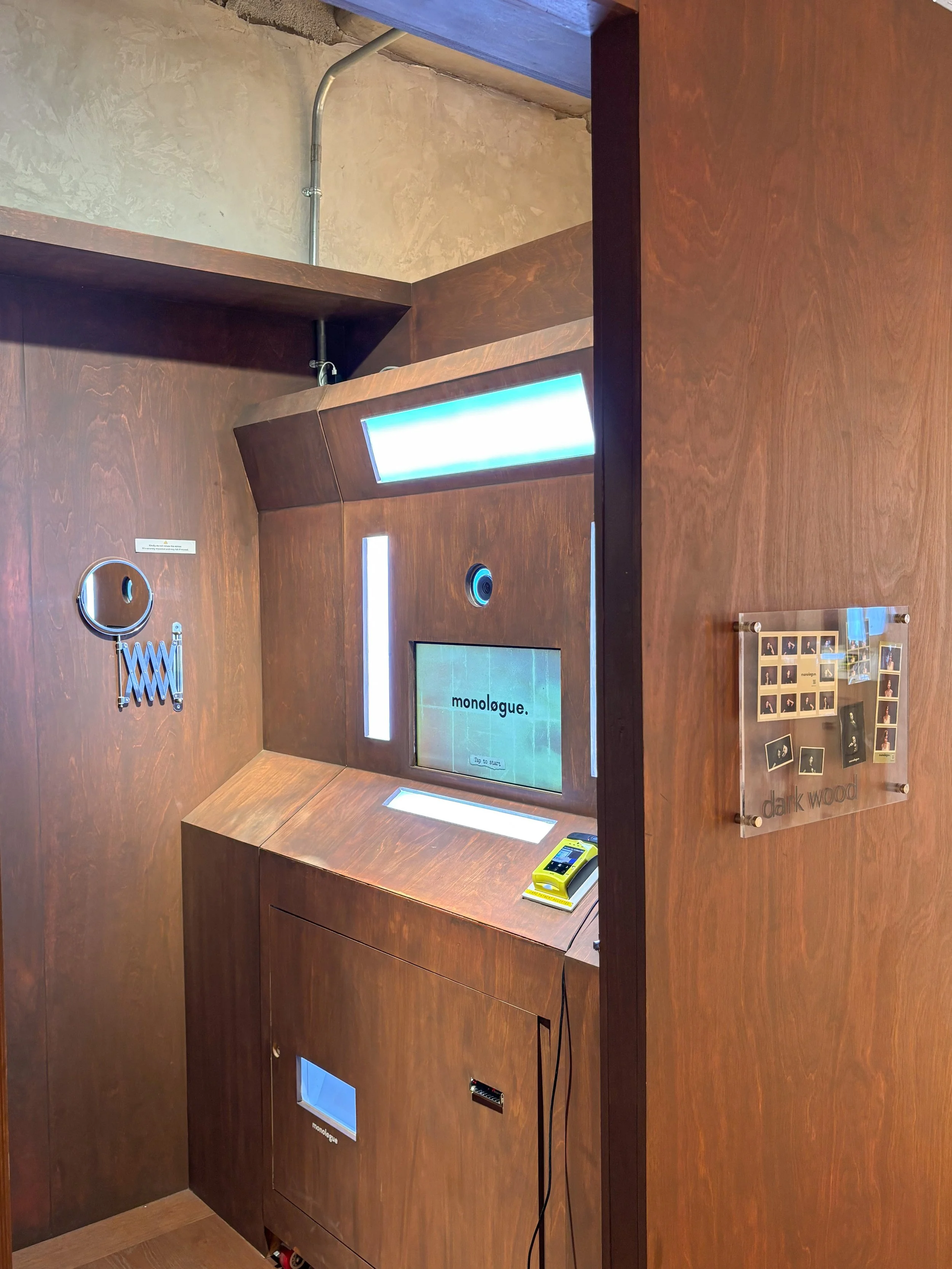 An interactive exhibit or installation with a screen displaying the word 'monologue.' on a wooden booth. The booth has light panels and objects like a mirror, a yellow device, and a small display case on the wooden surfaces. The background features a textured beige wall and a section of a blue ceiling.