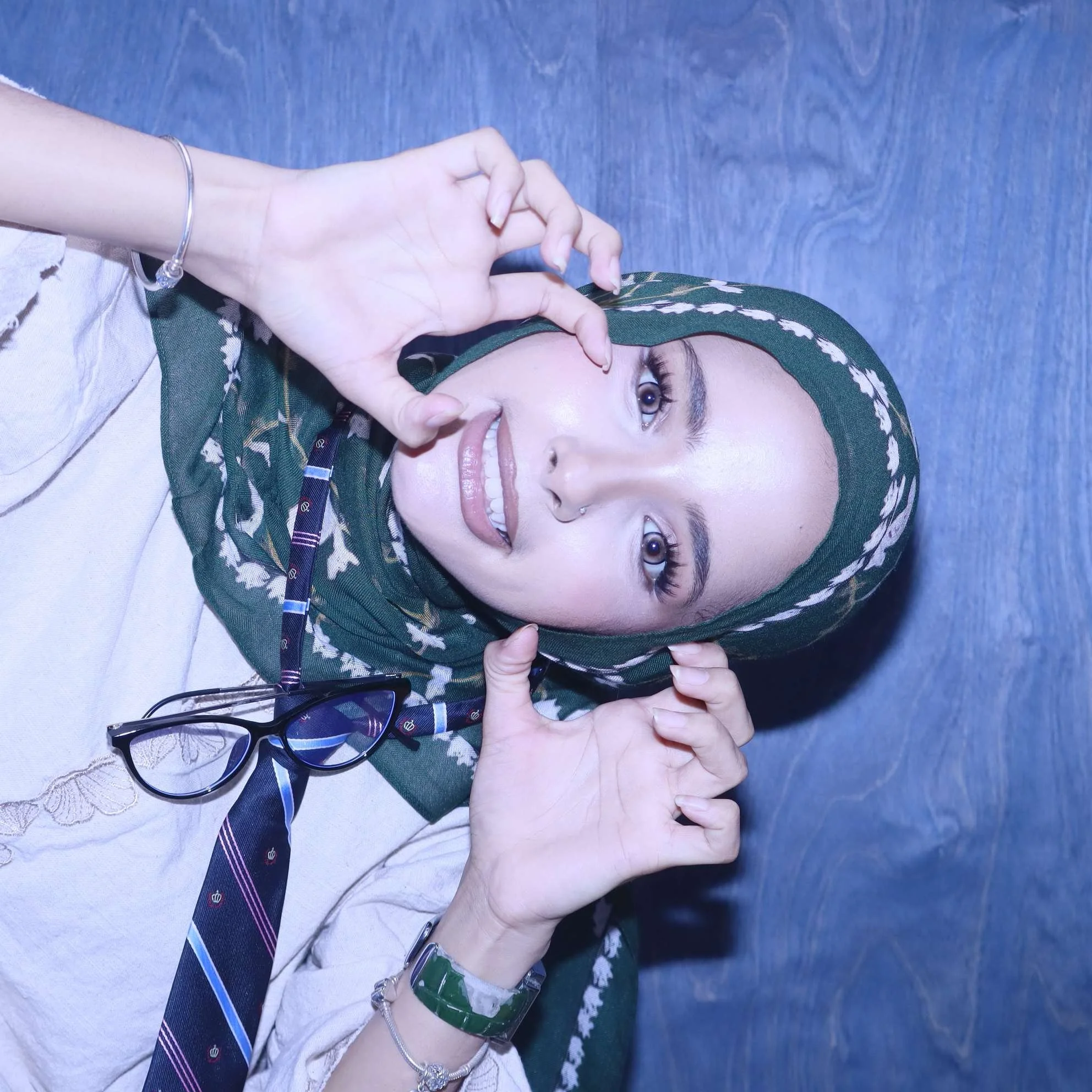 A woman wearing a green headscarf and beige dress making a heart shape with her hands, smiling, with glasses and jewelry on a blue background.