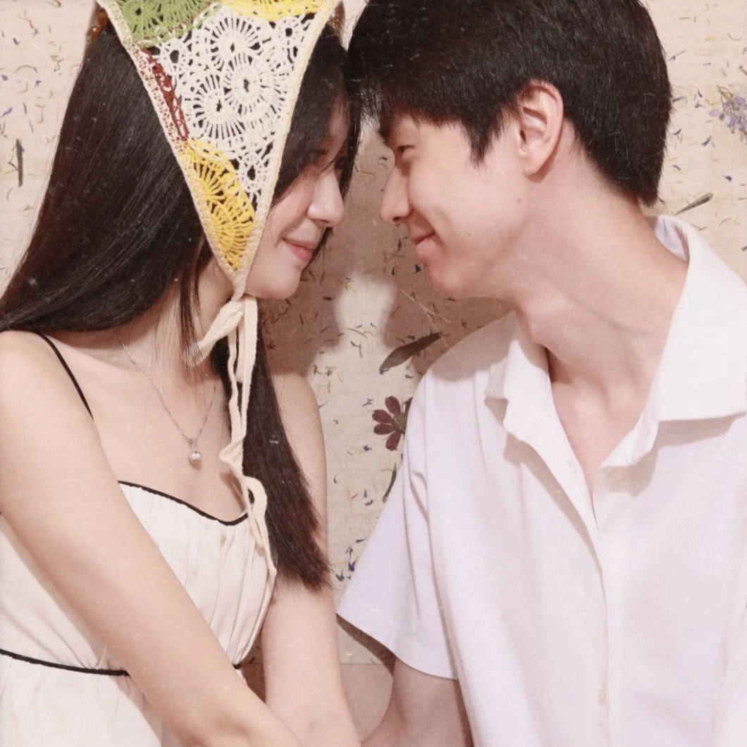 A young couple is touching foreheads, smiling, and gazing into each other's eyes indoors, with a floral wallpaper background.