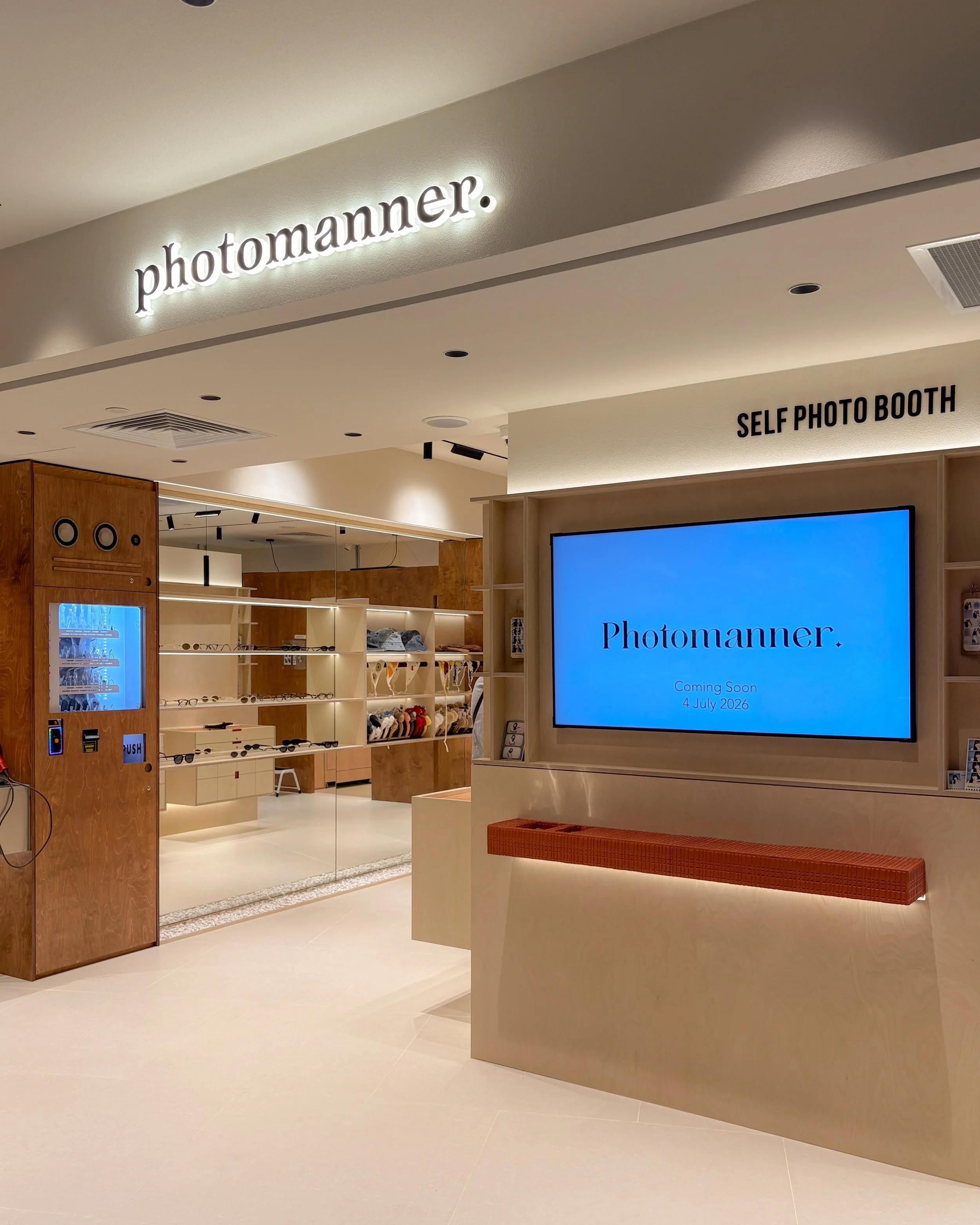 Interior of a modern retail store named 'photomanner' with a sign illuminated on the wall, a digital display showing the store name and opening date, and shelves with accessories in the background.