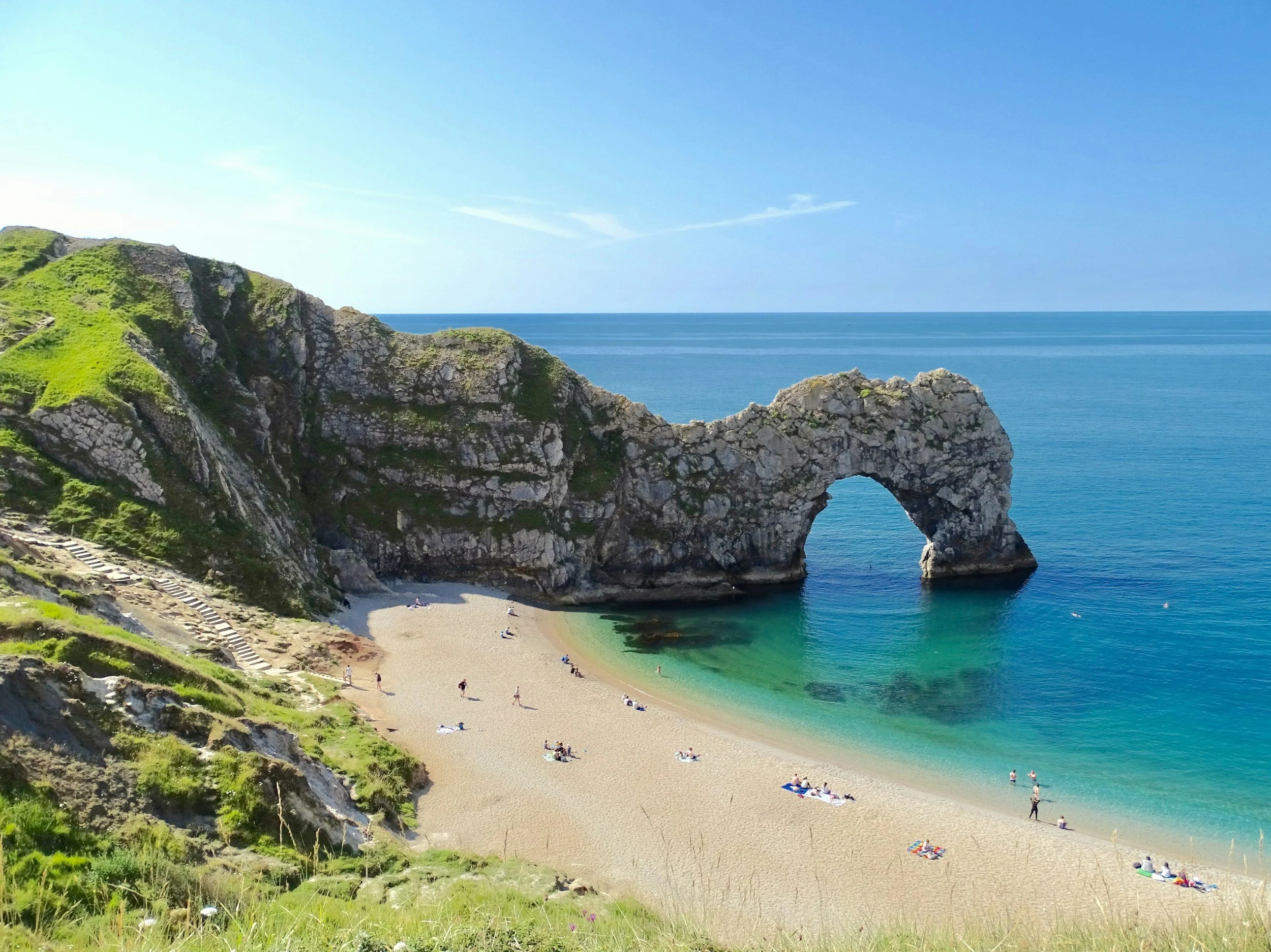 A coastal landscape featuring a large natural rock arch formation extending into the sea, with a sandy beach and people relaxing under a clear blue sky.