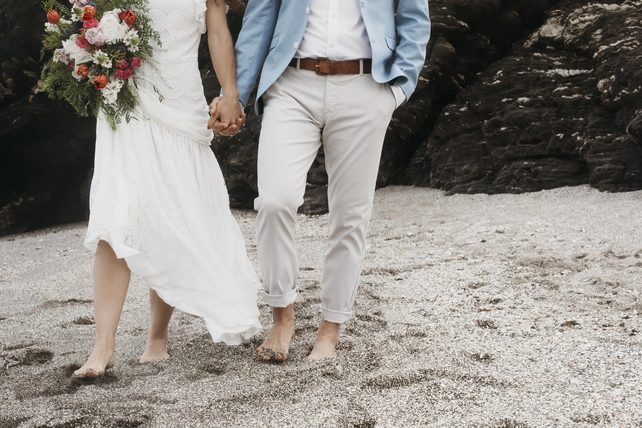 A couple holding hands while walking barefoot on a sandy beach, with the woman in a white lace dress and the man in light-colored trousers and a blue blazer, with dark rocks in the background.