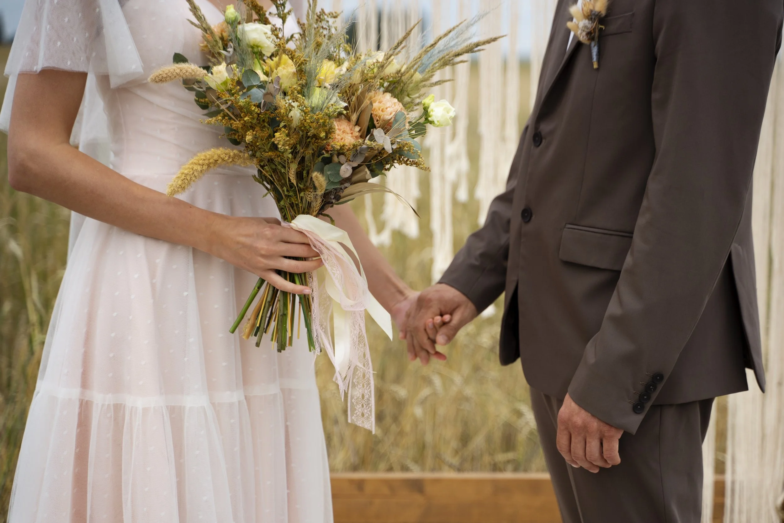 A bride holding a bouquet and a groom holding her hand during a wedding ceremony outdoors.