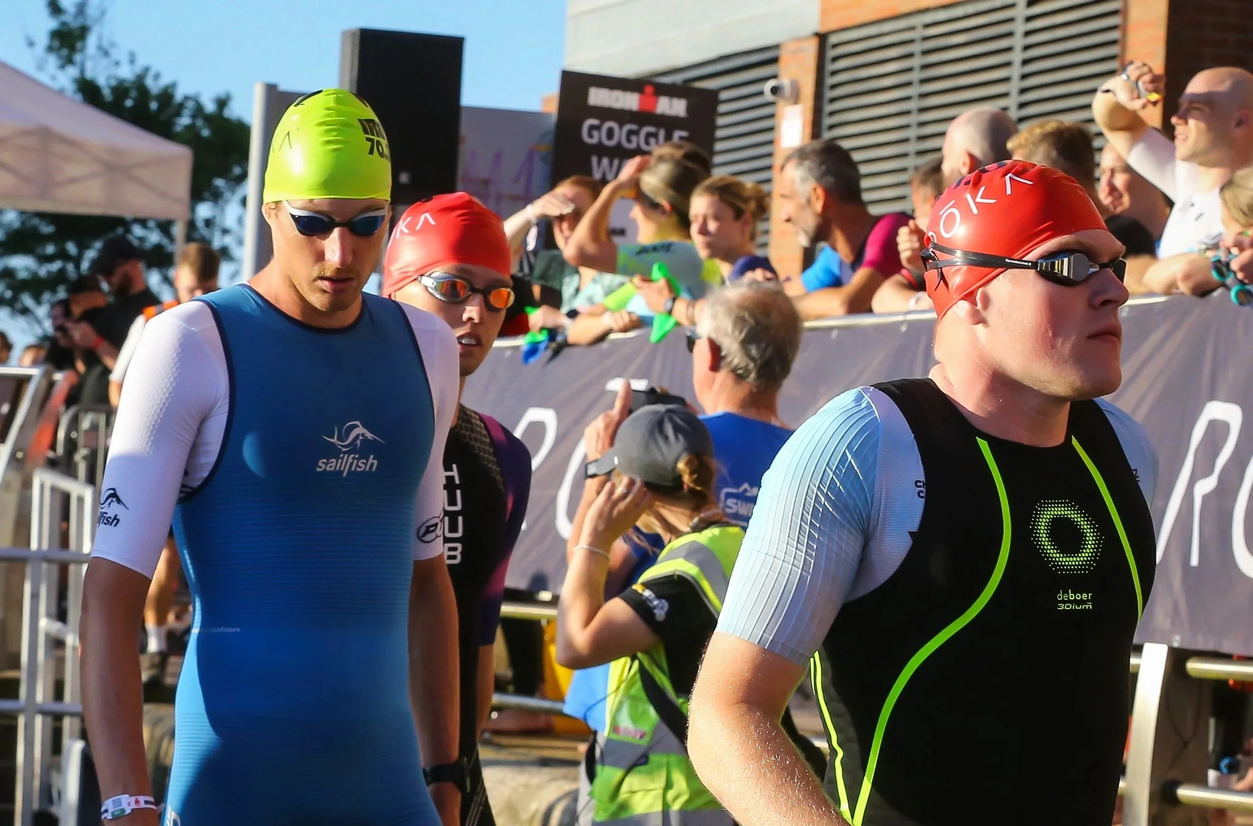 Triathletes wearing swim caps and goggles preparing for the race, with spectators and event signage in the background.
