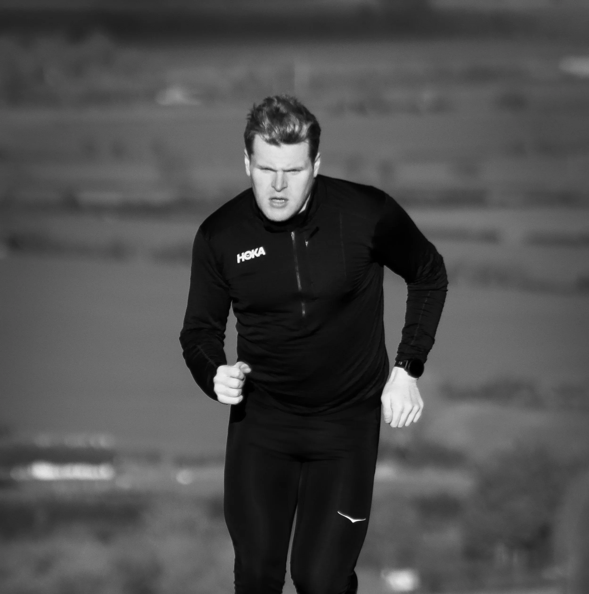 A black-and-white photo of a man running outdoors, wearing a dark athletic jacket with the brand name 'HOKA' on it.