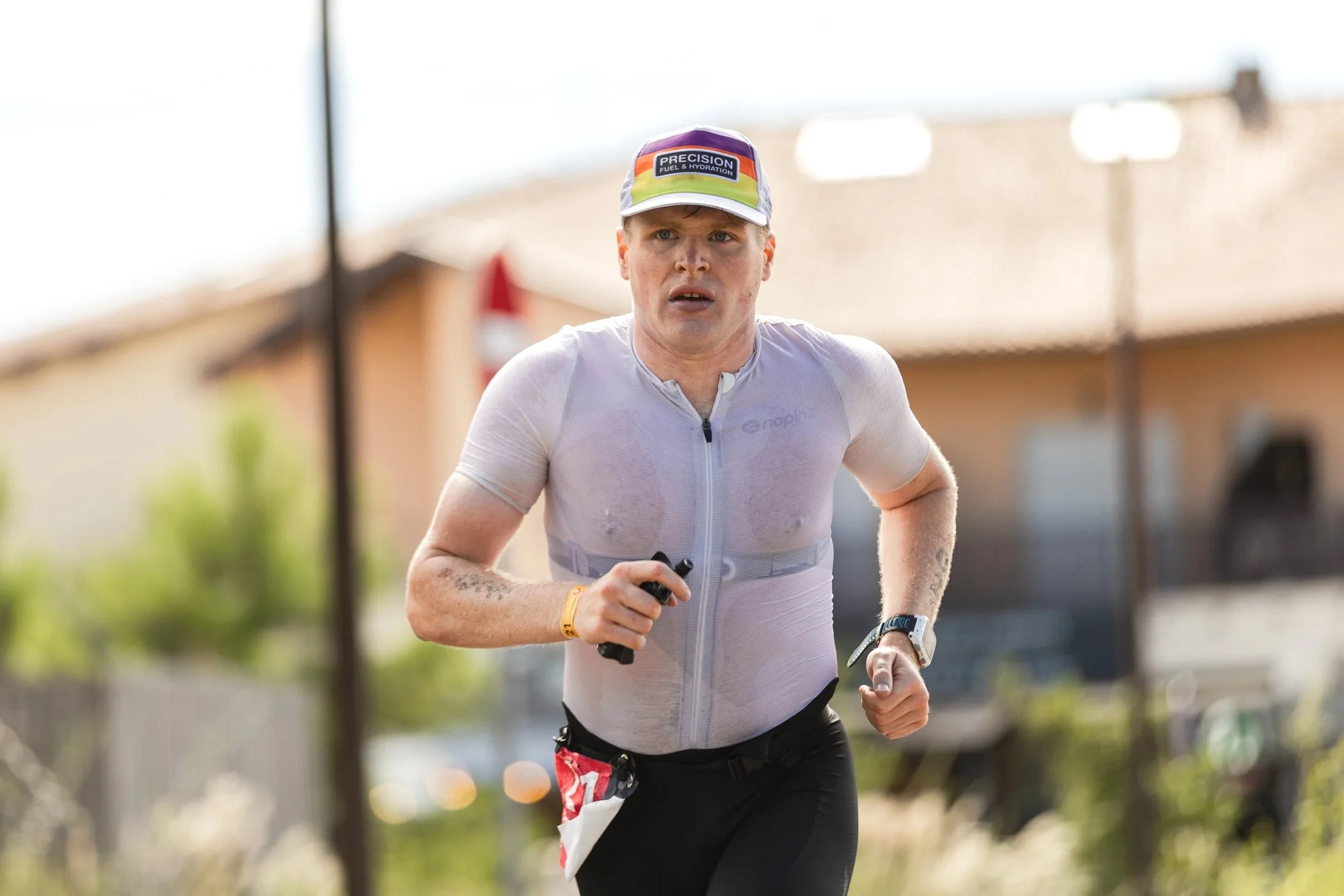 A male runner during a race, wearing a hat that says 'Precision Fuel & Hydration,' a wet, fitted shirt, black pants, and a wristwatch, running outdoors.