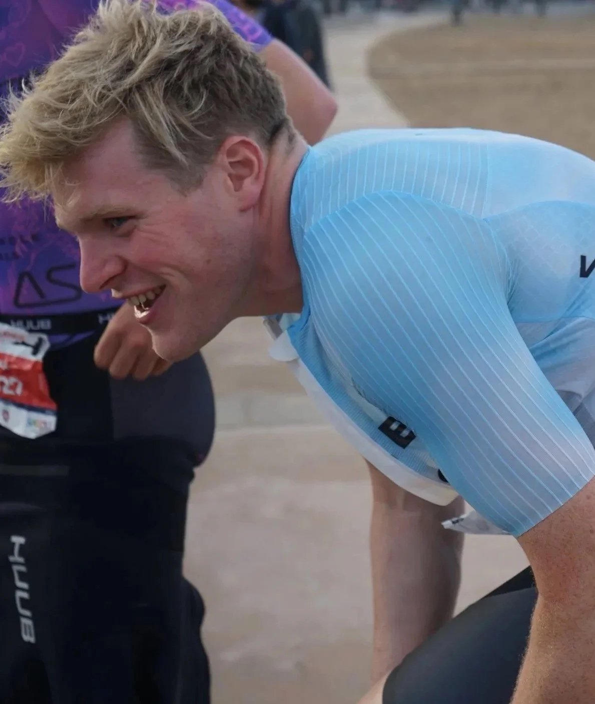 A male cyclist in a light blue jersey leaning forward and smiling during a race or event.