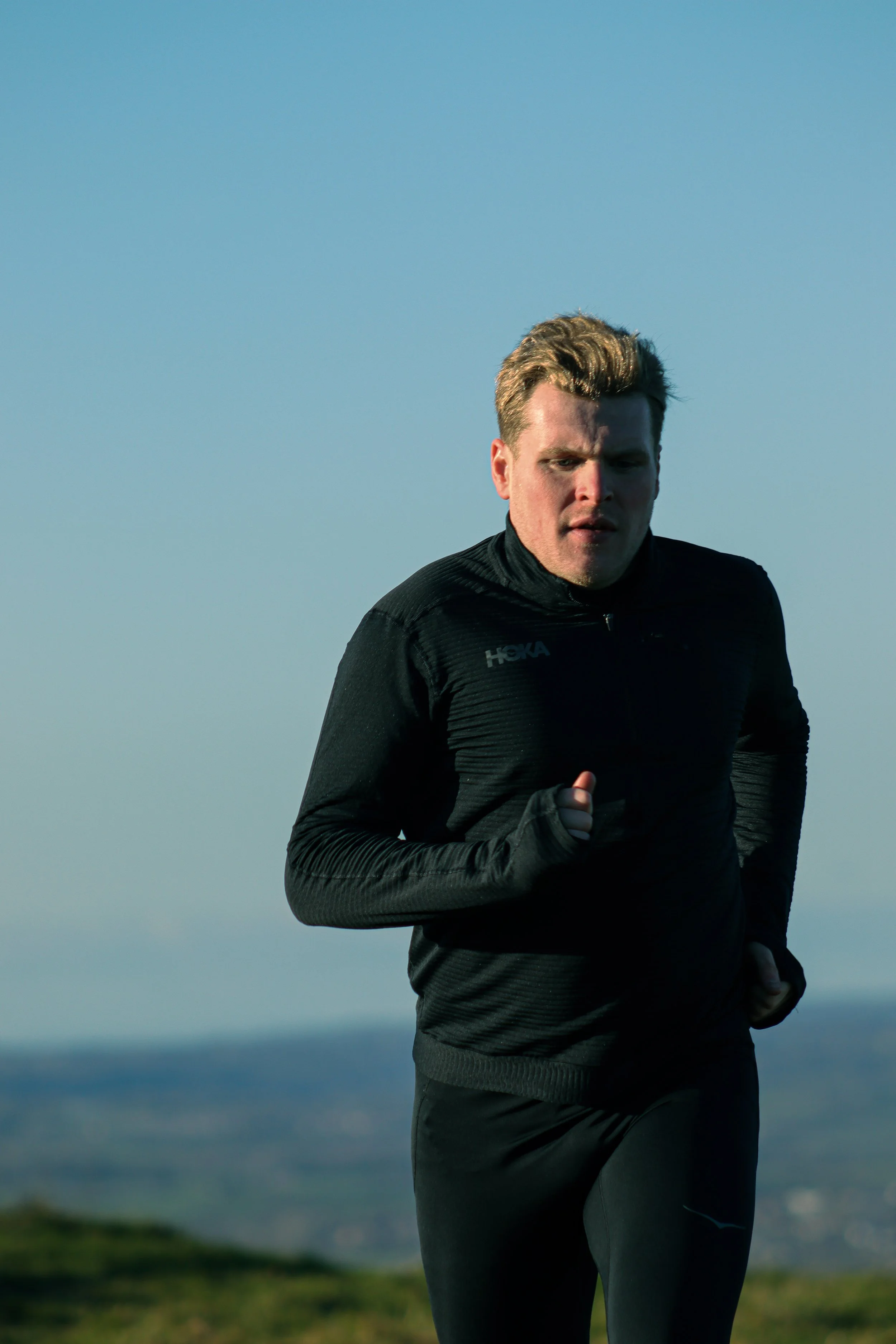 Man running outdoors on a cloudy day, wearing a black HOKA jacket and gray pants.