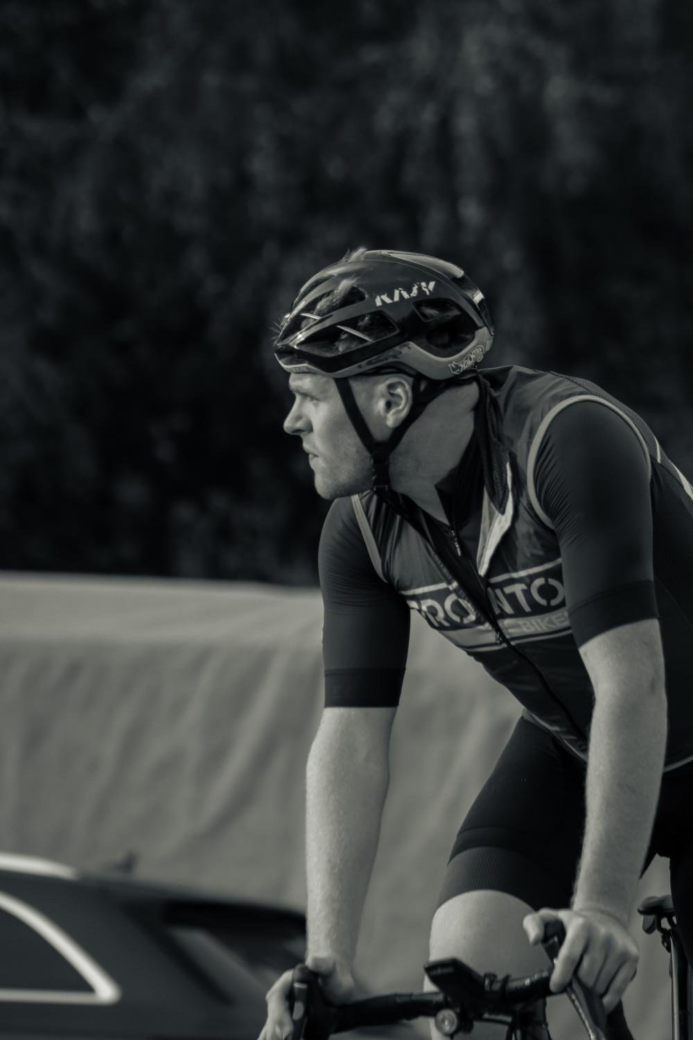 A man riding a bicycle, wearing a helmet and cycling gear, with a focused expression, on a road with a blurred background. why a coach is important for beginners and pro's