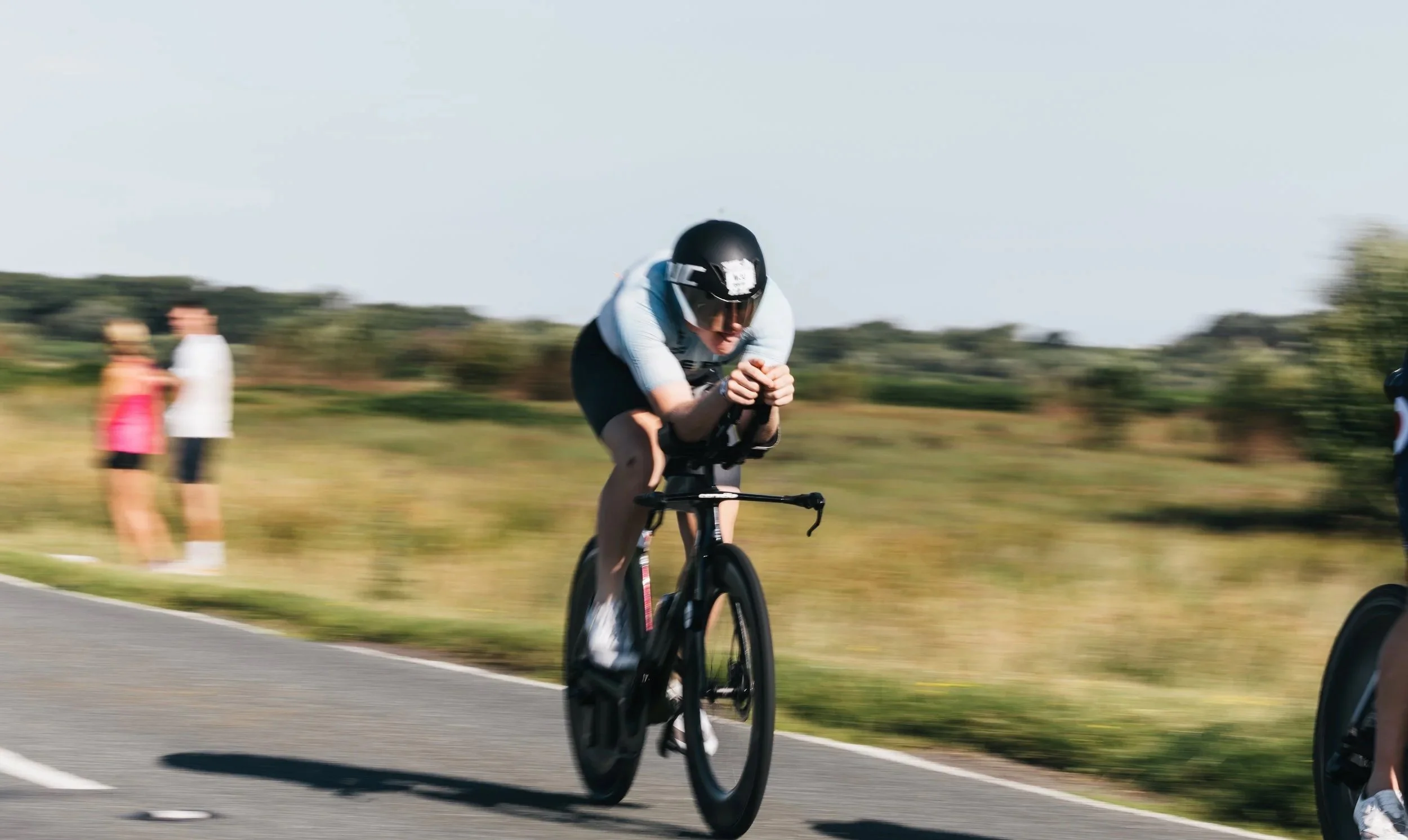 British Pro triathlete - A cyclist in a time trial race riding a bike on a paved road, wearing a helmet and aerodynamic clothing, with a grassy landscape in the background.