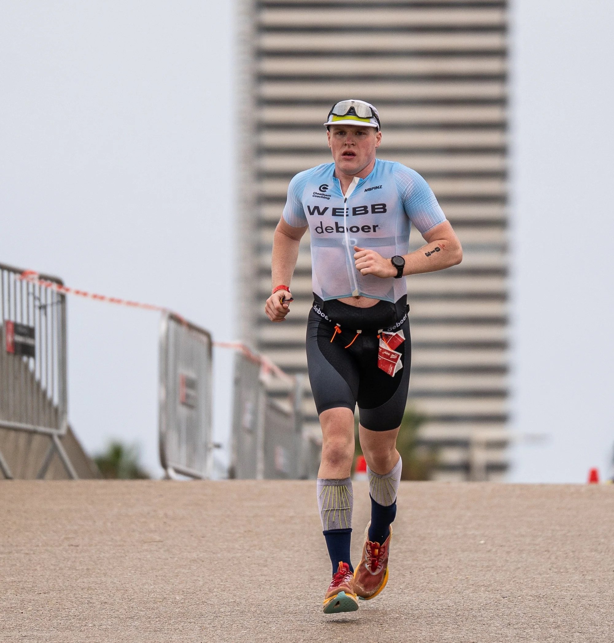 A male athlete running on a track during a race, wearing a light blue jersey, black shorts, and red running shoes, with a pair of sunglasses on his head.