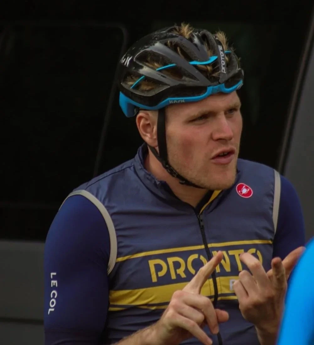 A male cyclist in a blue and yellow PRO-TOUR cycling jersey wearing a black and blue helmet, gesturing with his hands. Marathon to triathlon training