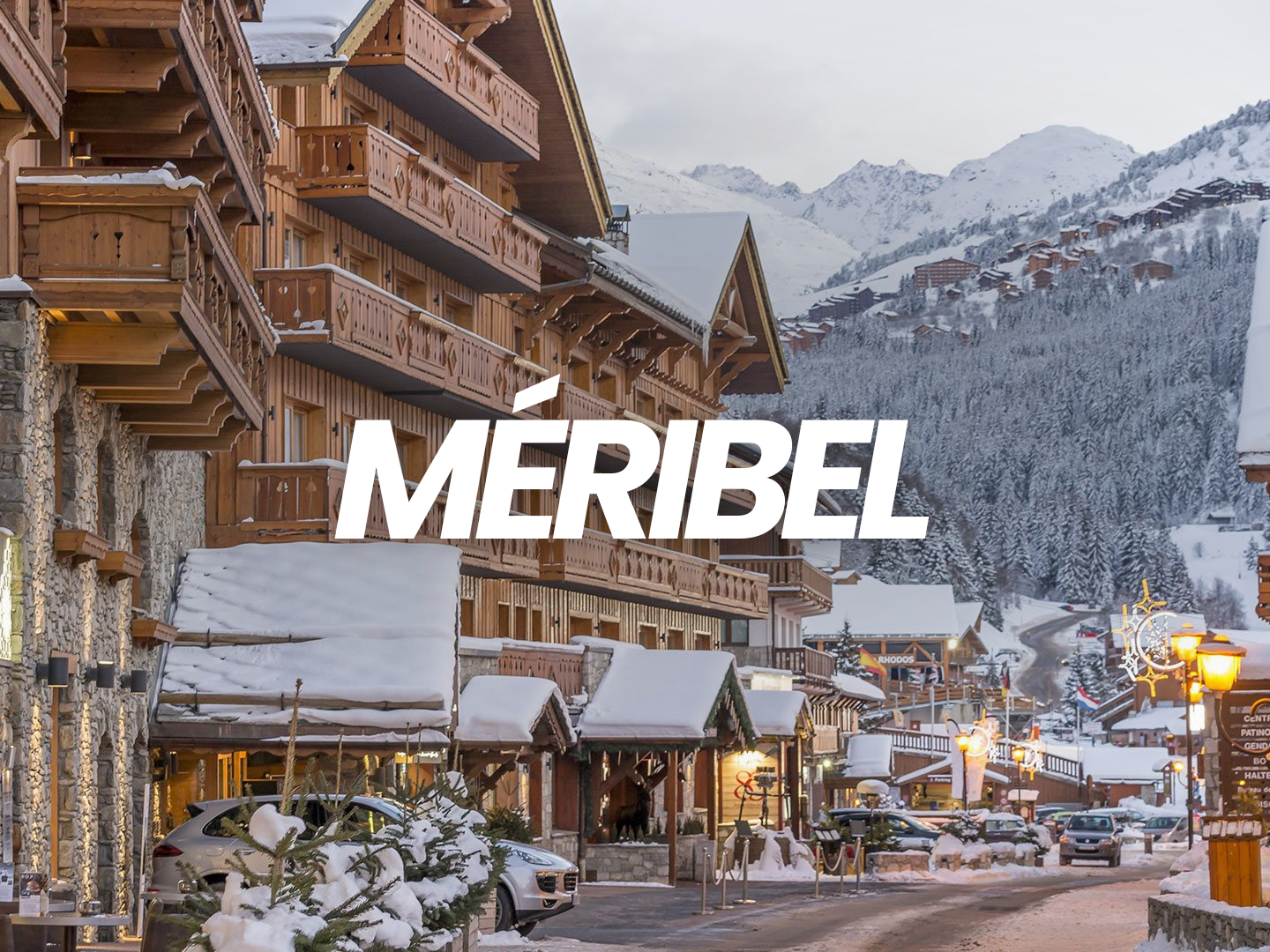 Ville alpine en hiver avec des bâtiments en bois sous la neige, montagnes en toile de fond, et des lampadaires allumés.