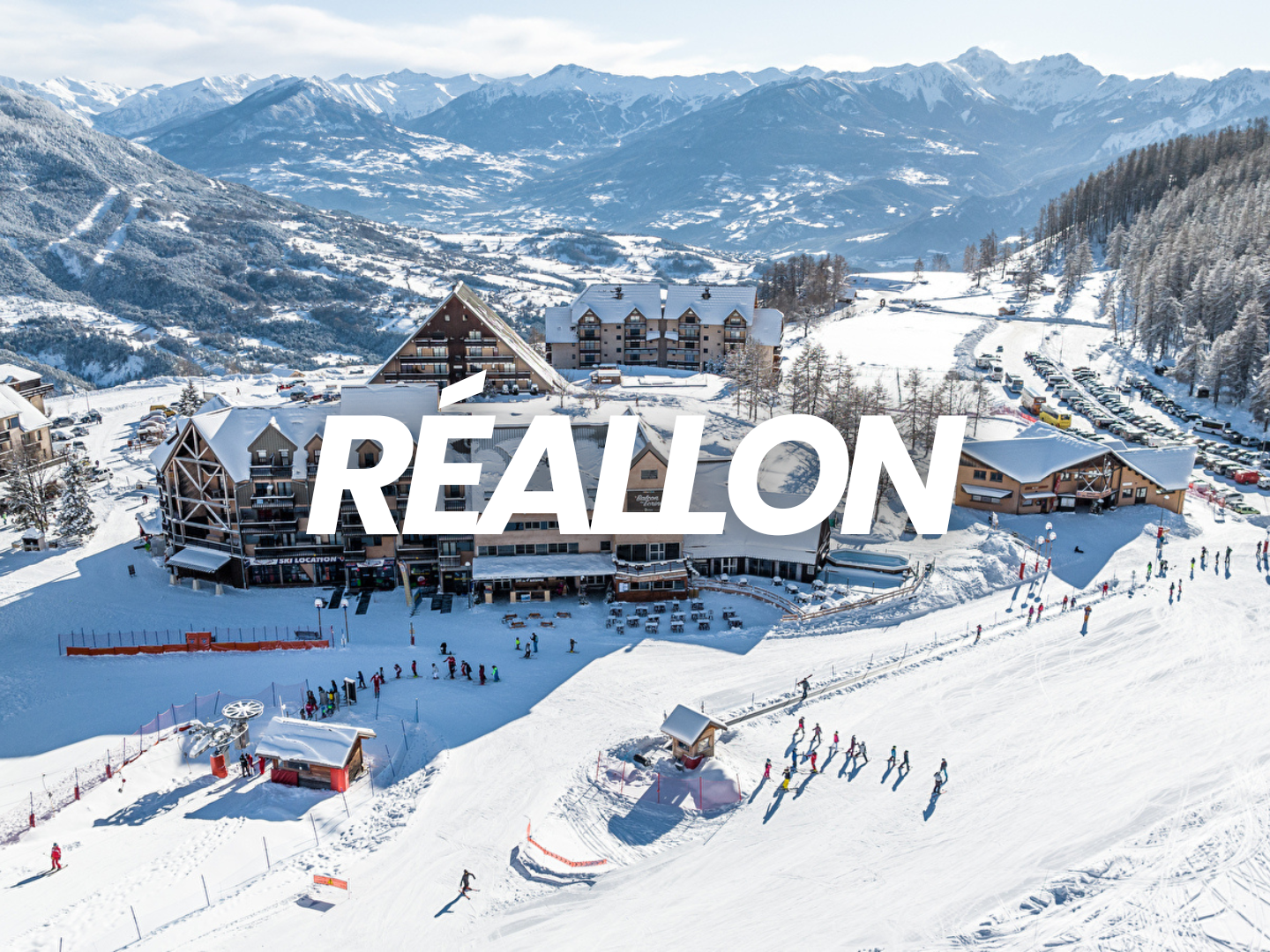 Vue d'une station de ski enneigée à Réallon, avec des bâtiments en bois, des skieurs et un paysage montagneux en arrière-plan.