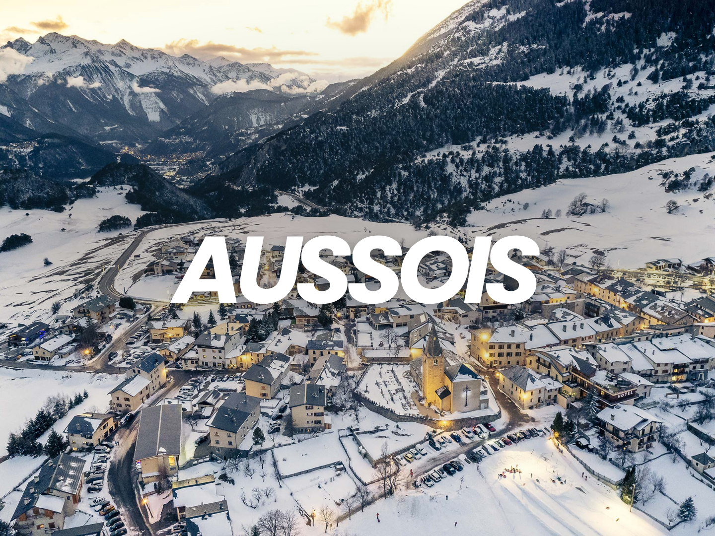 Vue aérienne d'un village enneigé à Aussois, avec montagnes en arrière-plan et éclairage nocturne dans quelques bâtiments.