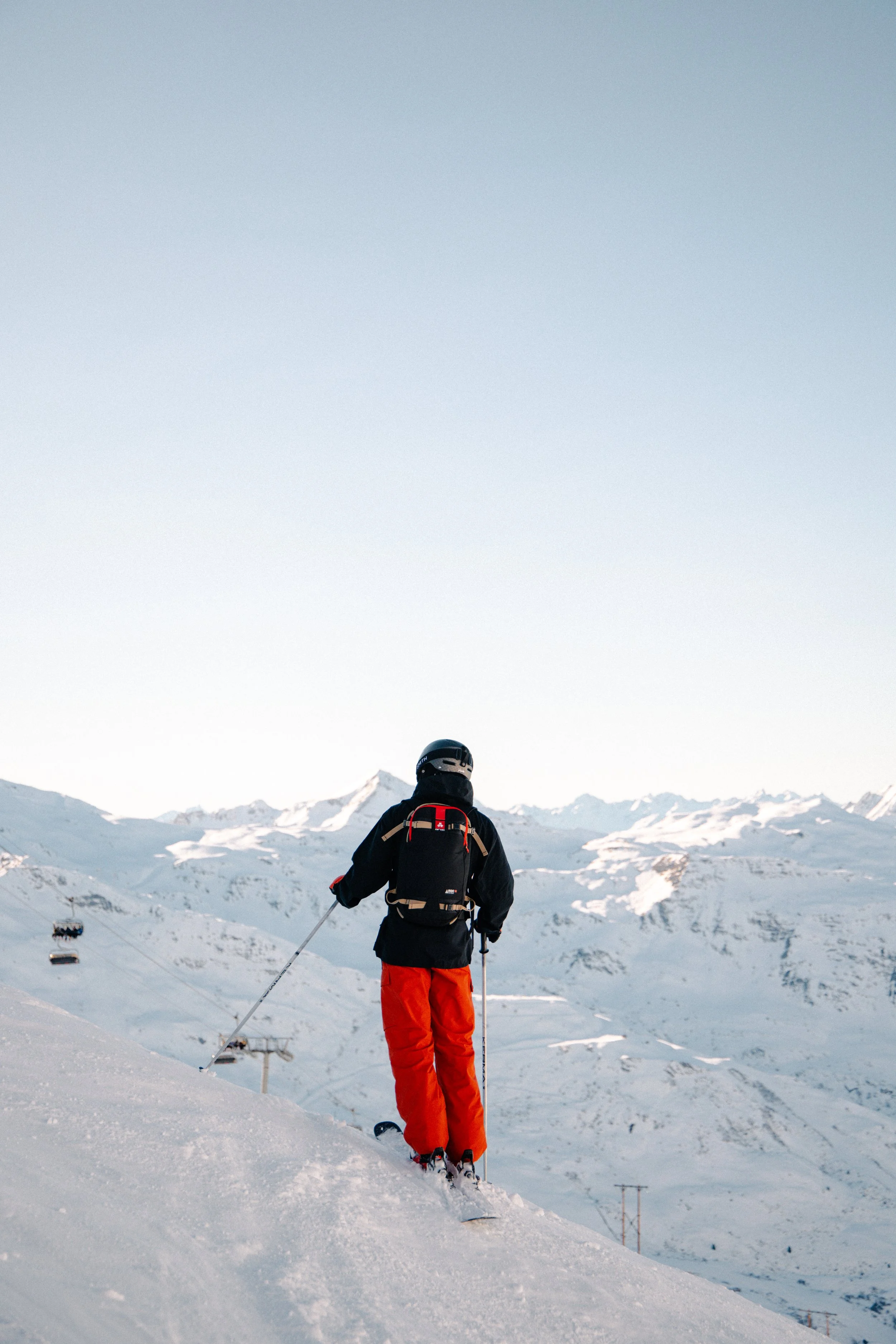 A skier dressed in black and red gear standing on snowy mountain slope, mountain range in background.
