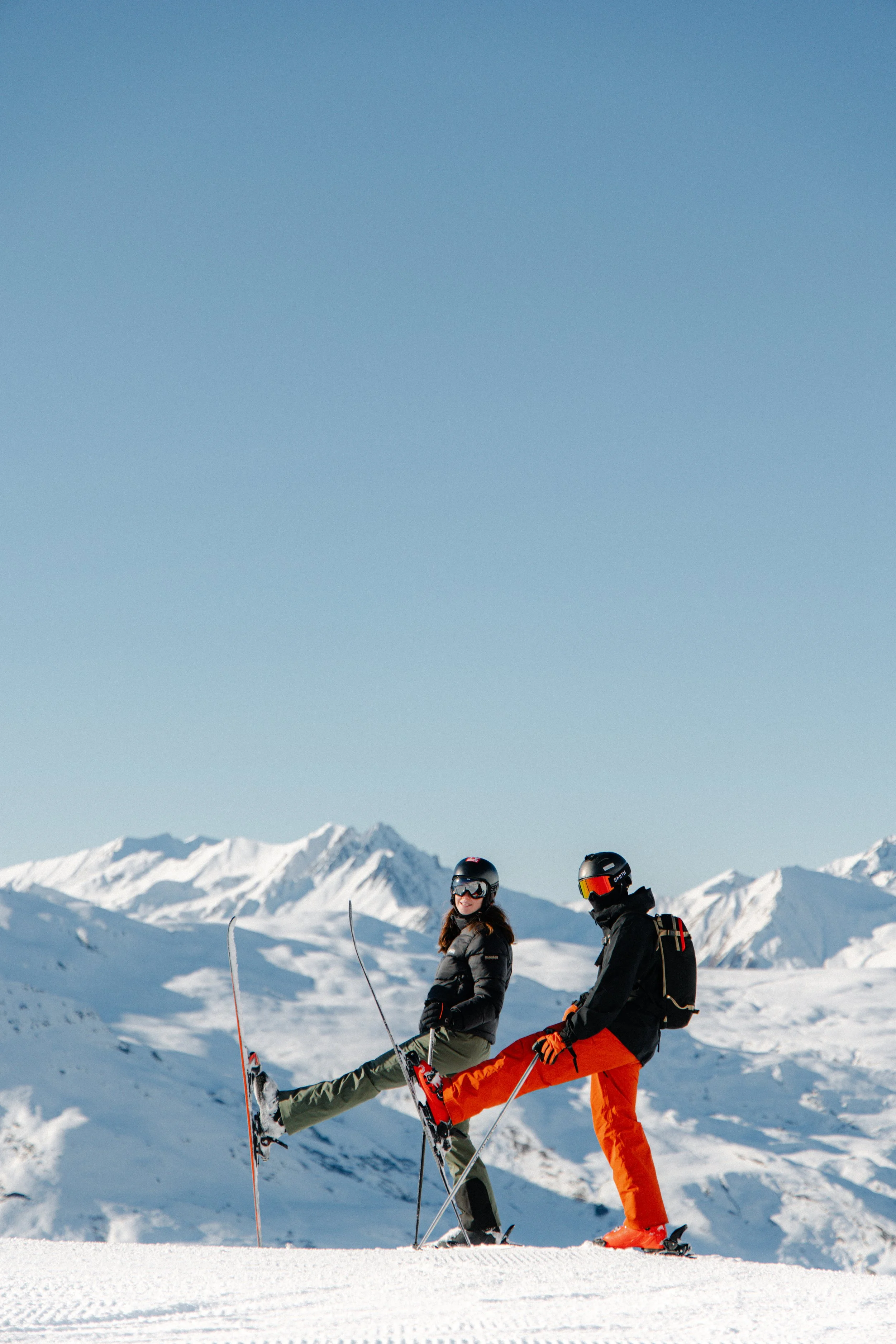 Two skiers in winter gear on a snowy mountain with clear blue sky and snow-covered peaks in the background.