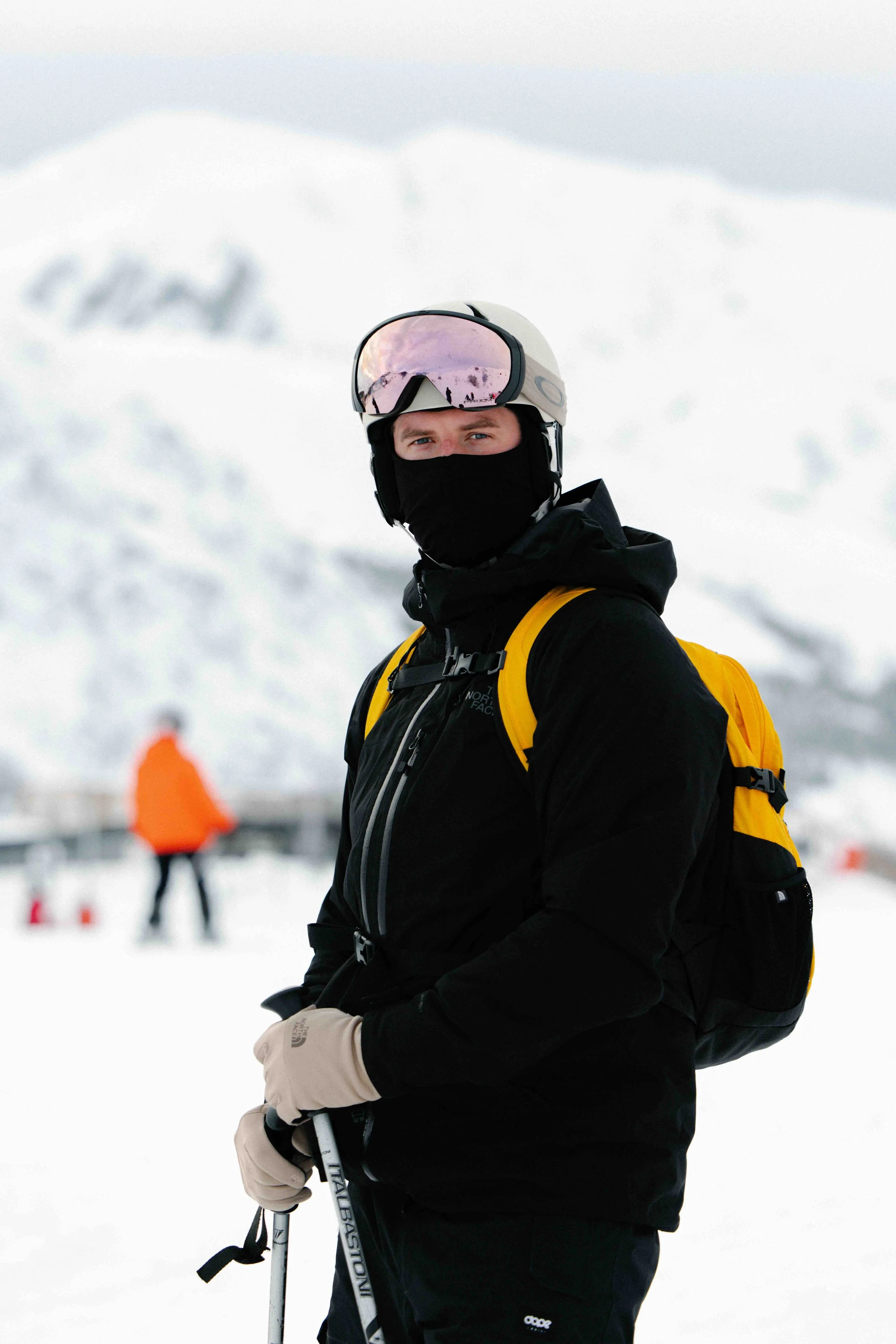 A man dressed in winter gear, including a helmet, goggles, a black jacket, gloves, and carrying a yellow backpack, standing in a snowy landscape with snow-covered mountains in the background.