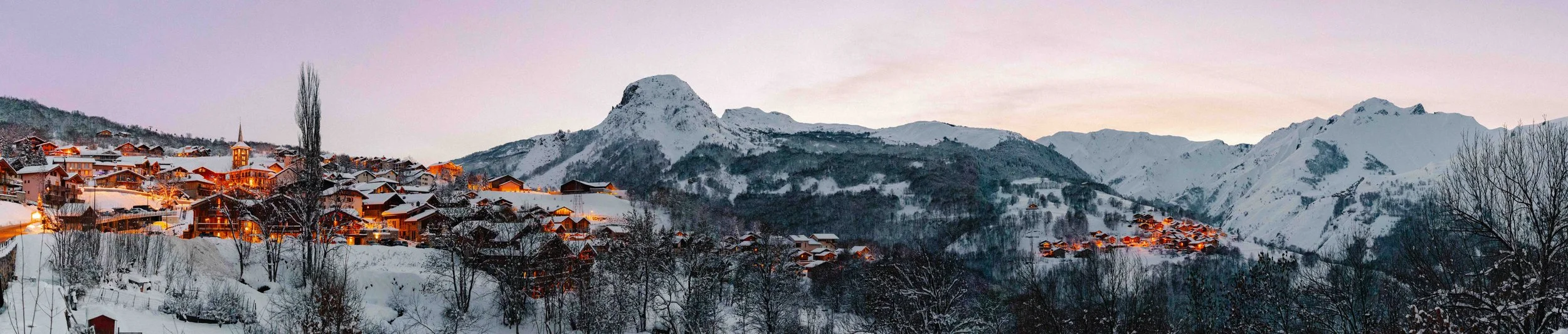 A snowy mountain village at dusk with snow-covered rooftops, bare trees, and mountains in the background.