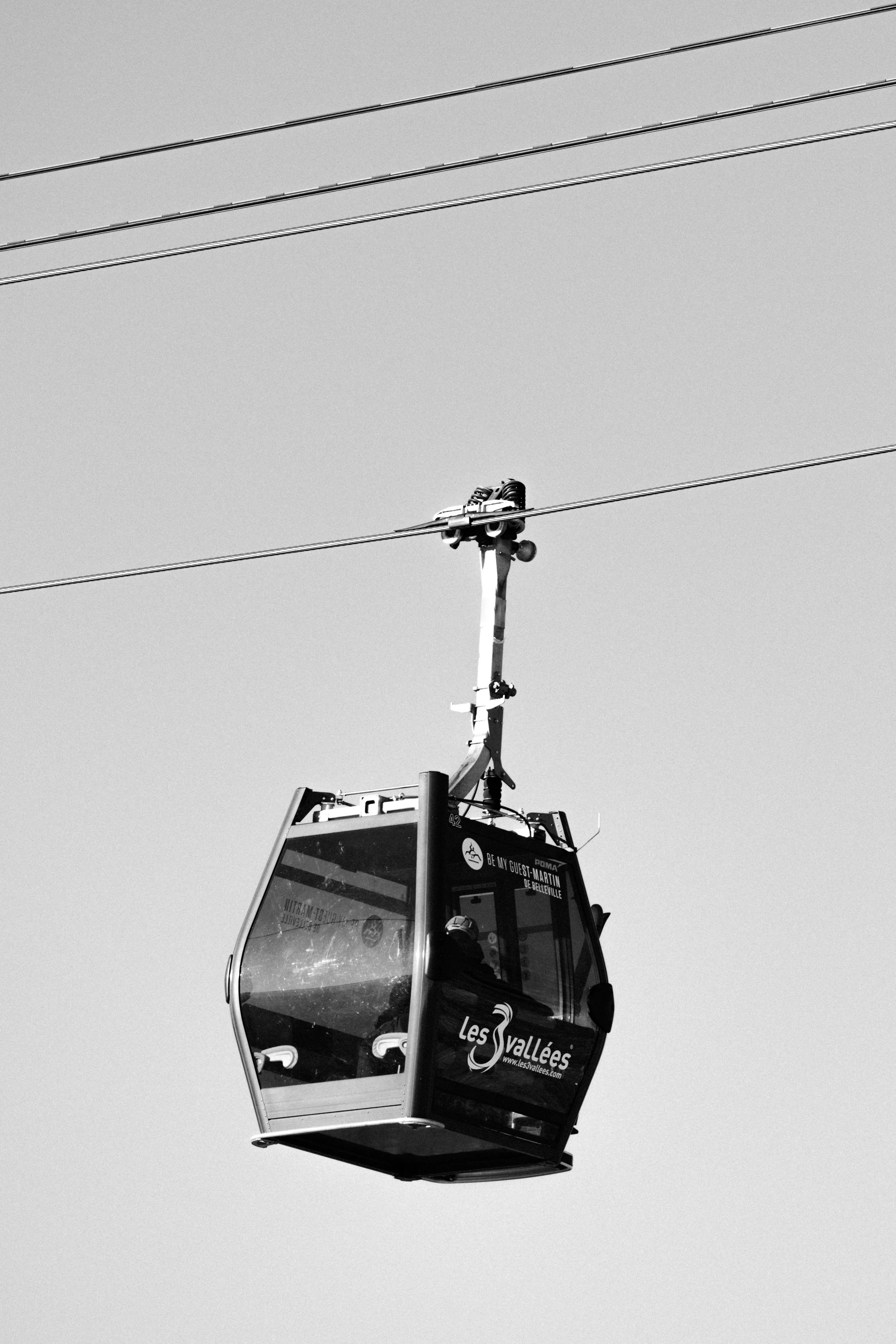 A black and white photo of a cable car suspended in mid-air, against a clear sky. The cable car has 'Les Vallées' written on it and is passing over power lines.