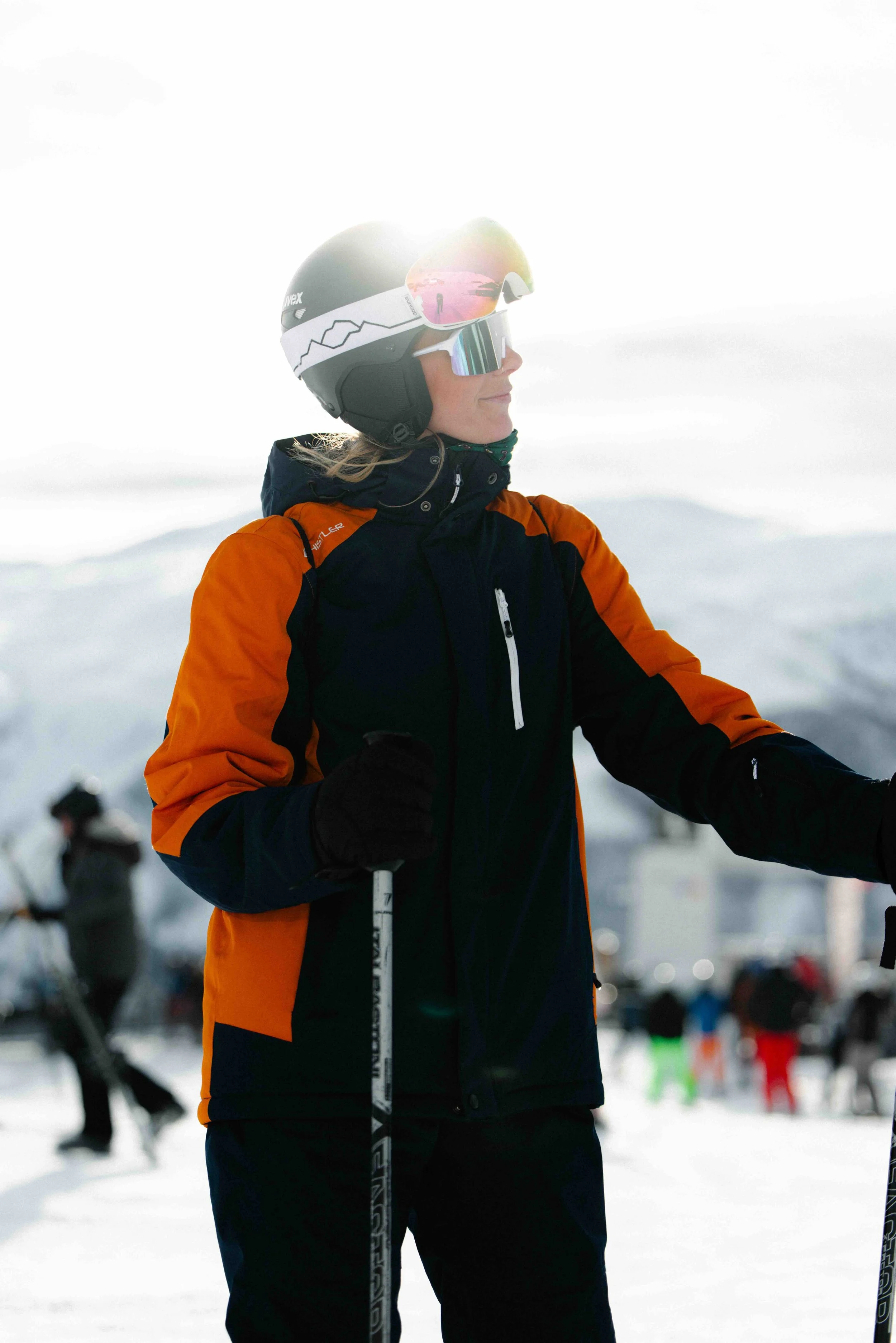 A woman in ski gear, including a helmet and goggles, standing outdoors in a snowy landscape with other skiers in the background.