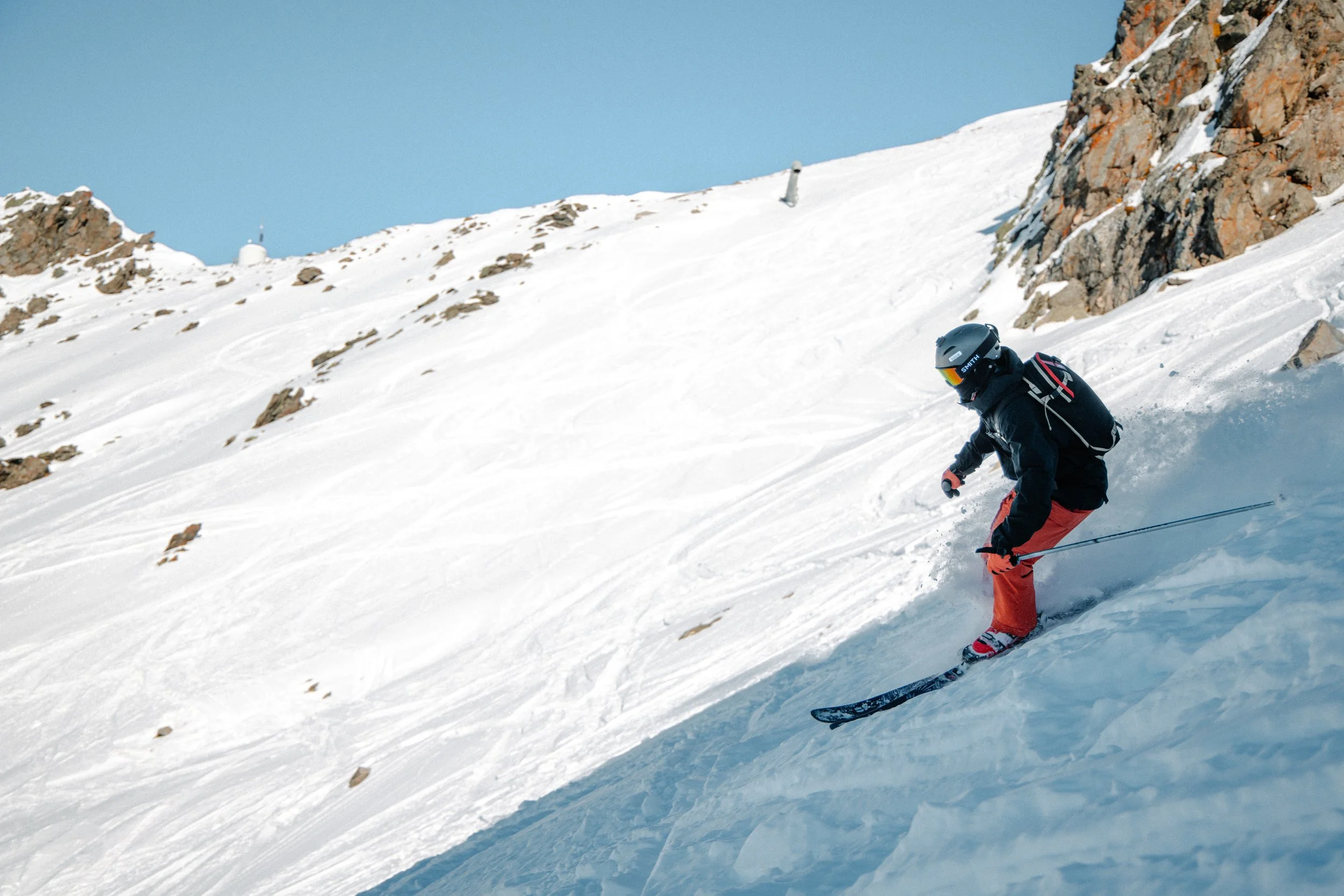 A person skiing down a snowy mountain slope, wearing a helmet, goggles, and winter gear under a clear blue sky.