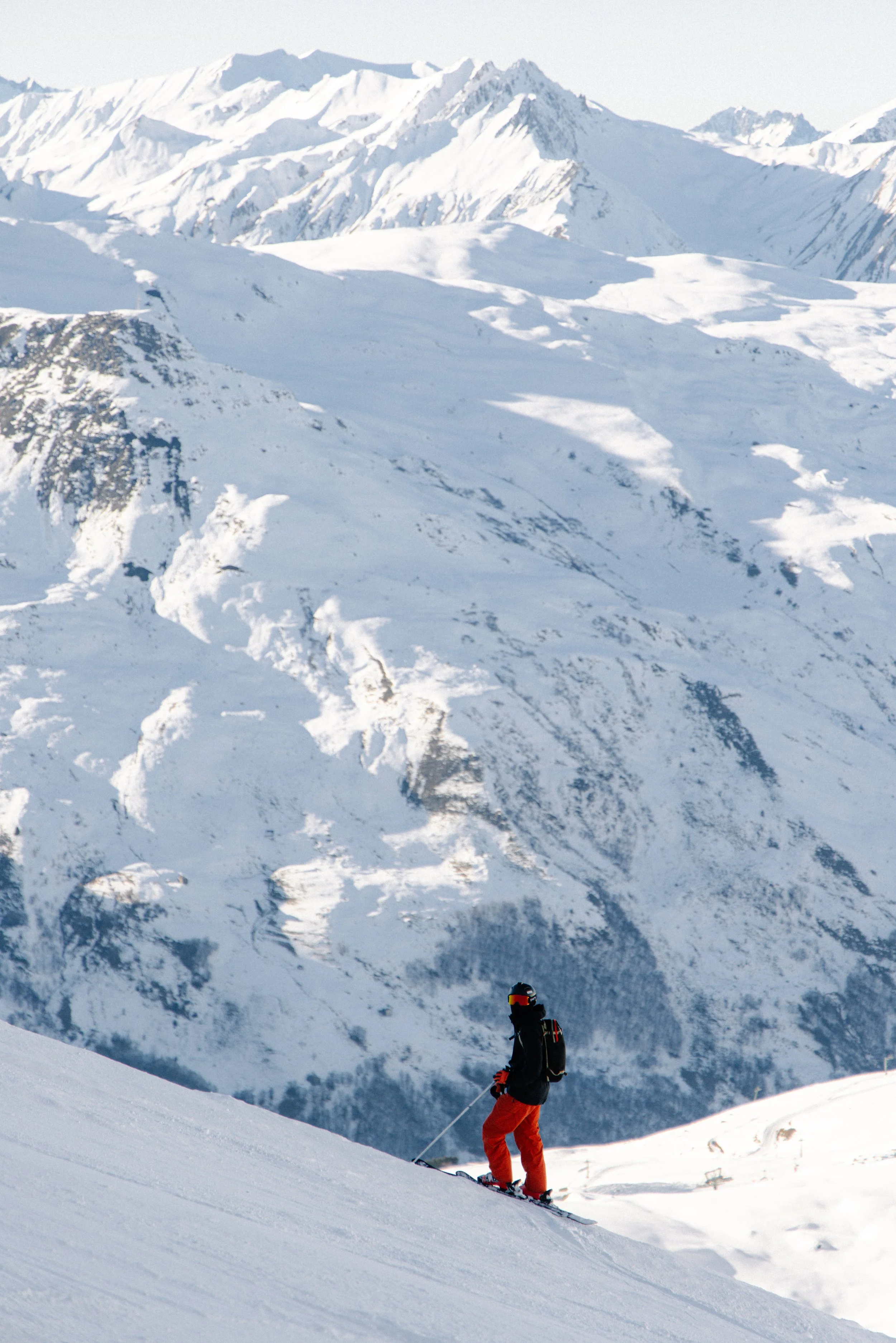 A skier dressed in black and orange gear skiing down a snowy mountain slope with tall snow-covered mountains in the background.