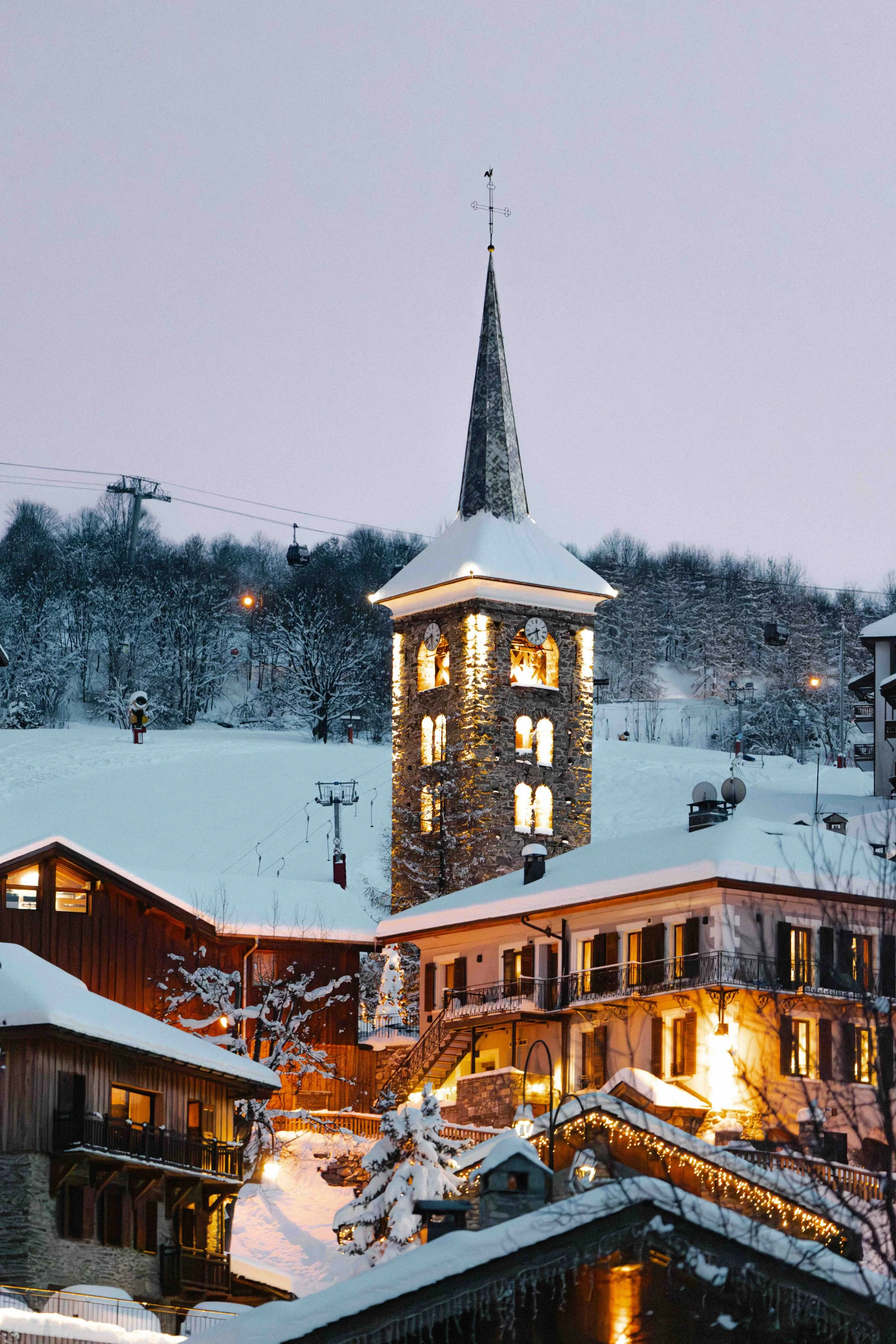 Snow-covered village with illuminated clock tower and buildings, evening scene with lights