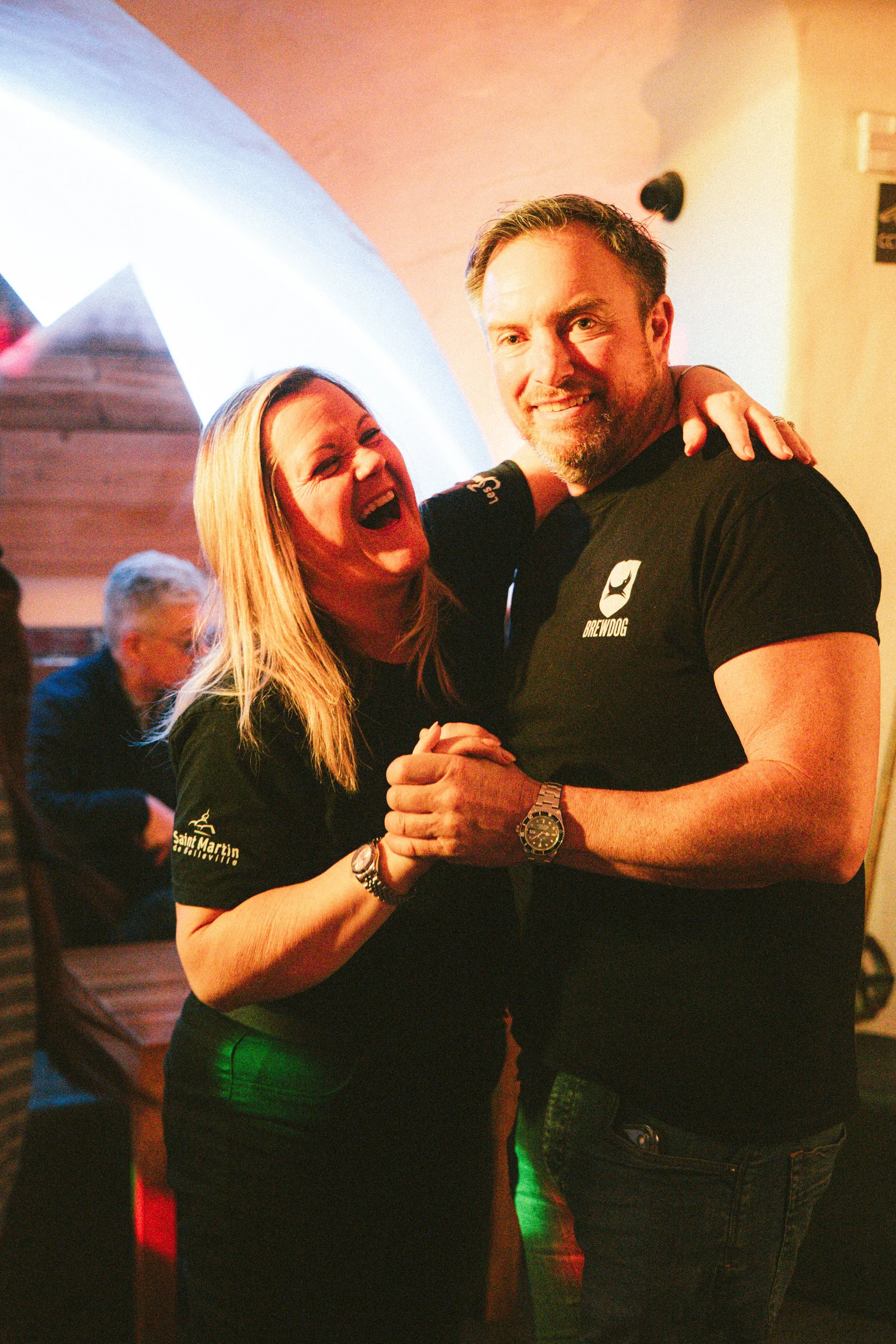 A couple dancing and smiling at a social gathering, with a background of other people and warm lighting.