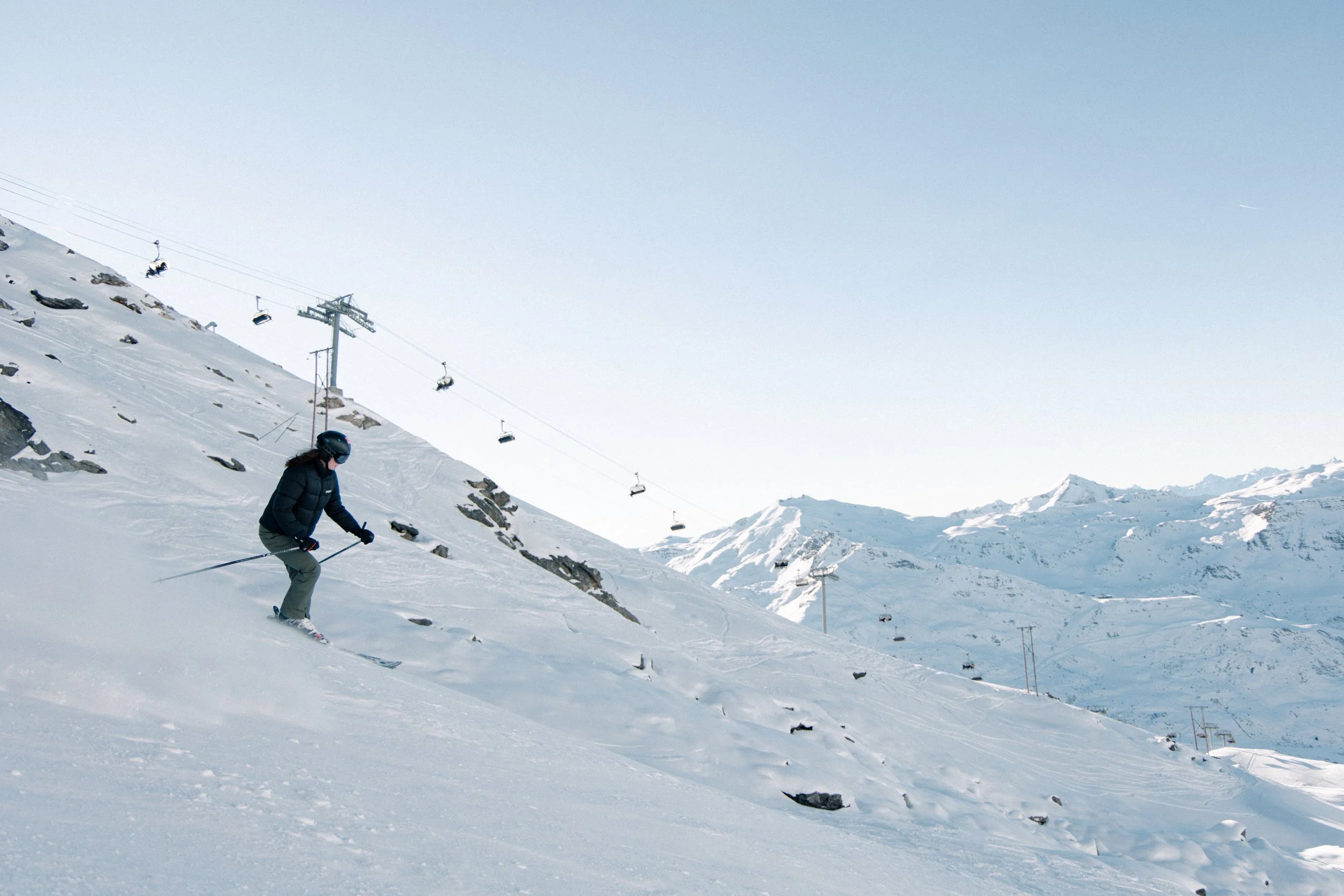 A person skiing downhill on a snowy mountain slope with ski lifts in the background and snow-covered mountain peaks under a clear sky.