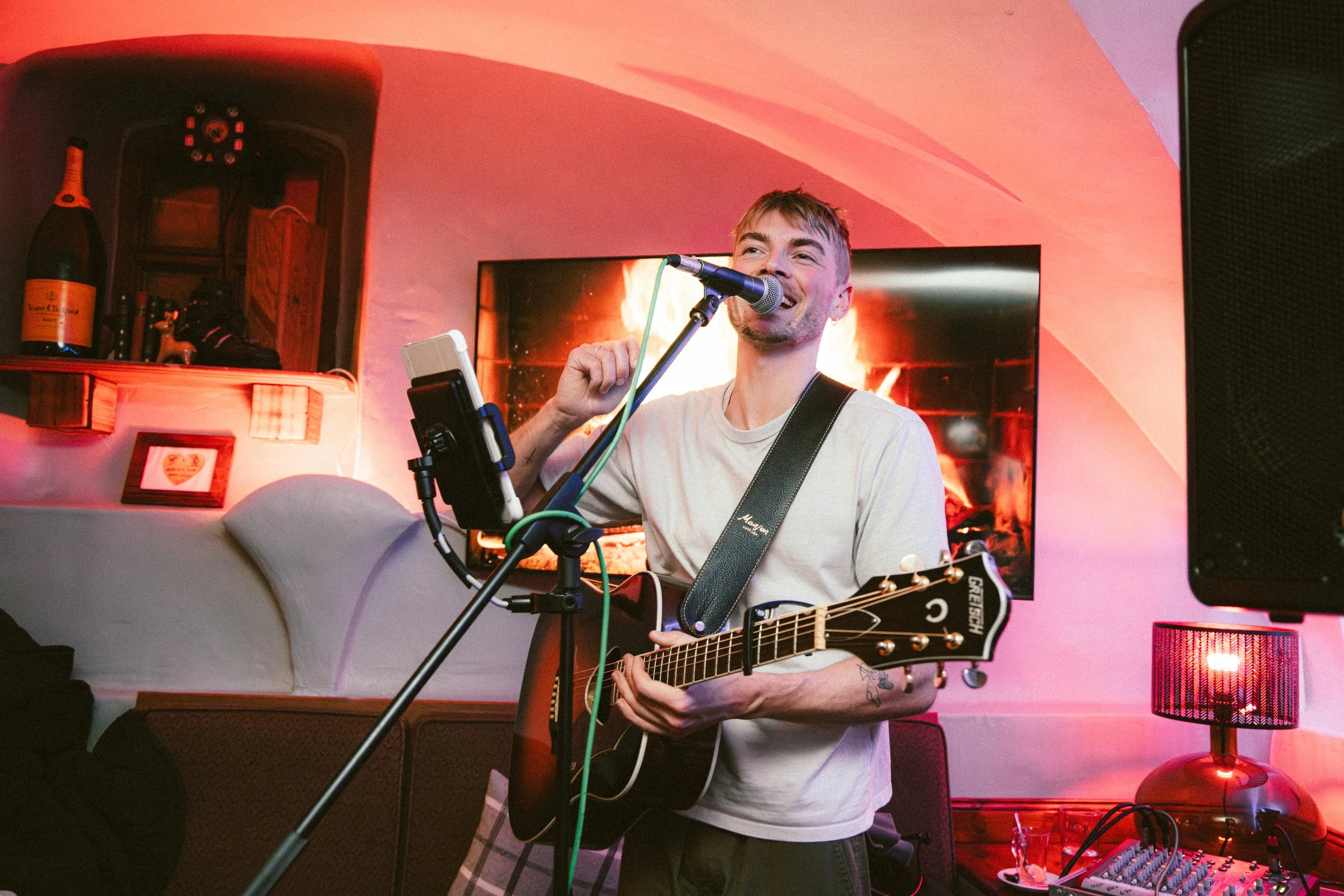 A man singing into a microphone while playing an acoustic guitar in a cozy room with pink and orange lighting, a fireplace on the wall behind him, and a sound mixer on a table to the right.