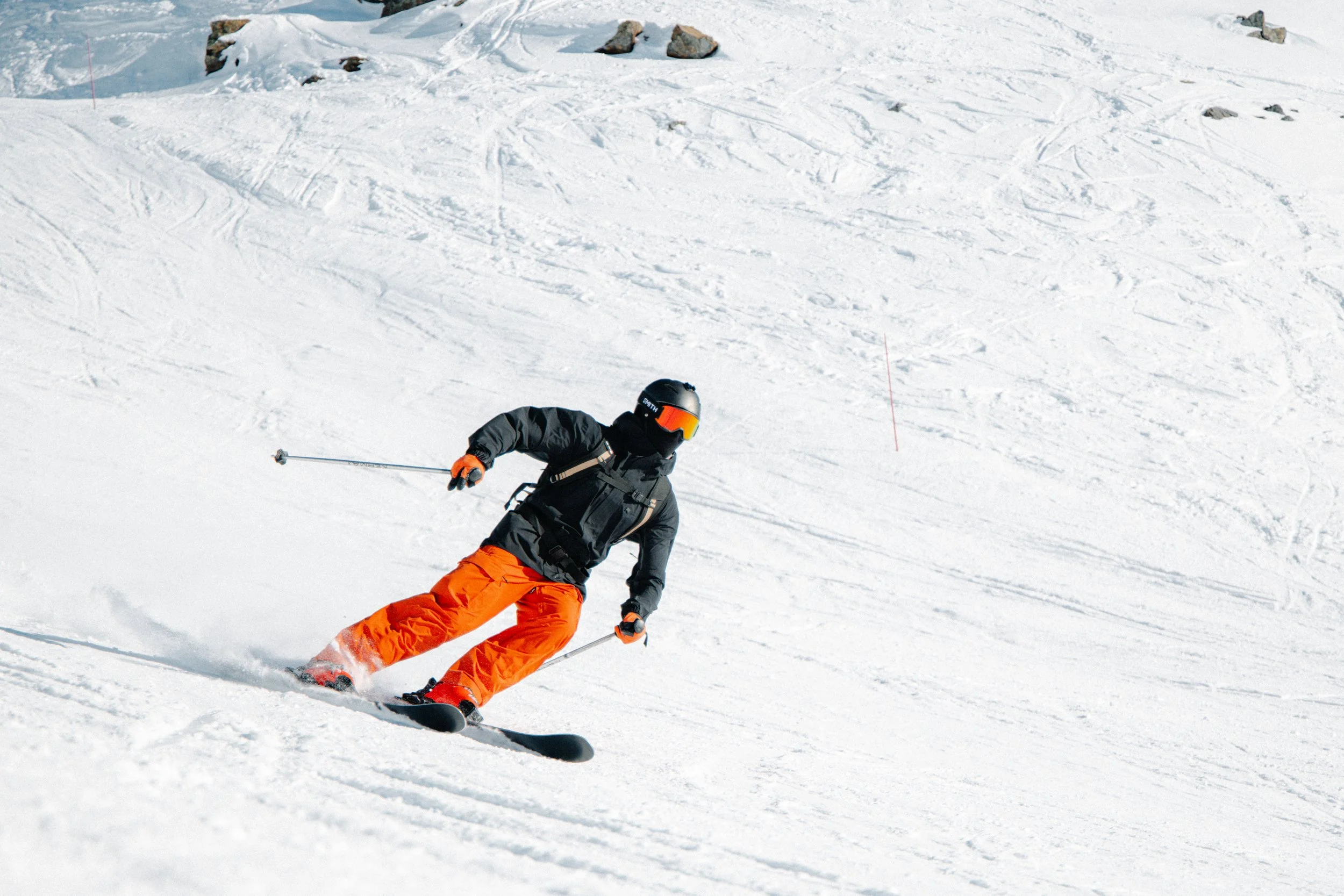 A skier in black jacket and orange pants skiing down a snowy slope with ski poles, wearing a helmet and goggles.