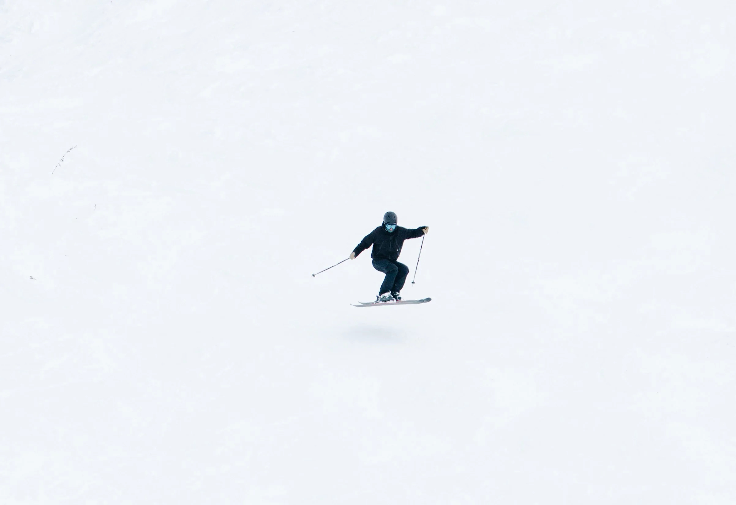 A person skiing downhill on snow, wearing a black helmet, black jacket, black pants, and goggles, with ski poles in both hands.
