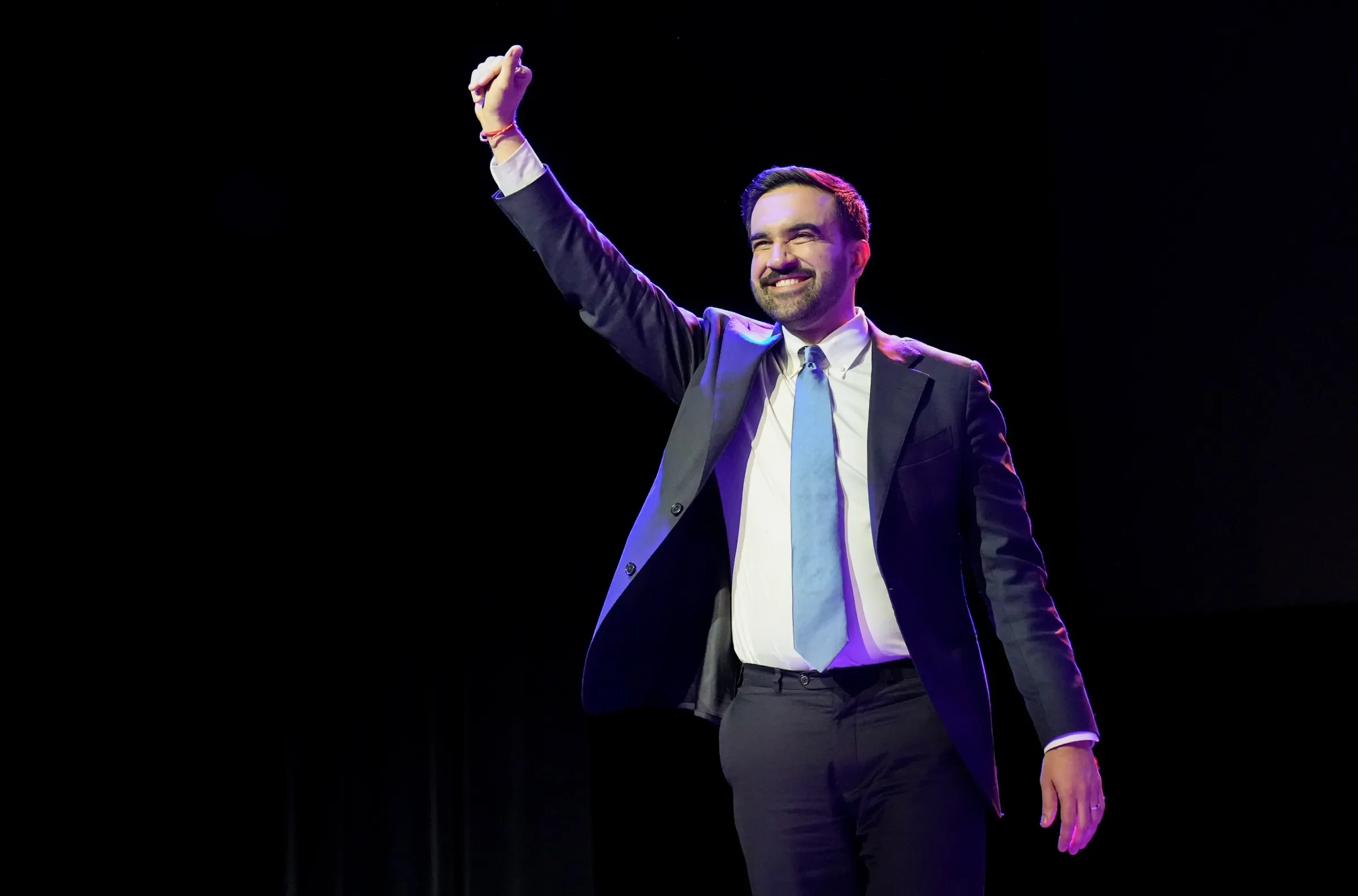 Siyah arka plana sahip sahnede, gülümseyen ve elini havaya kaldırmış genç bir adam.