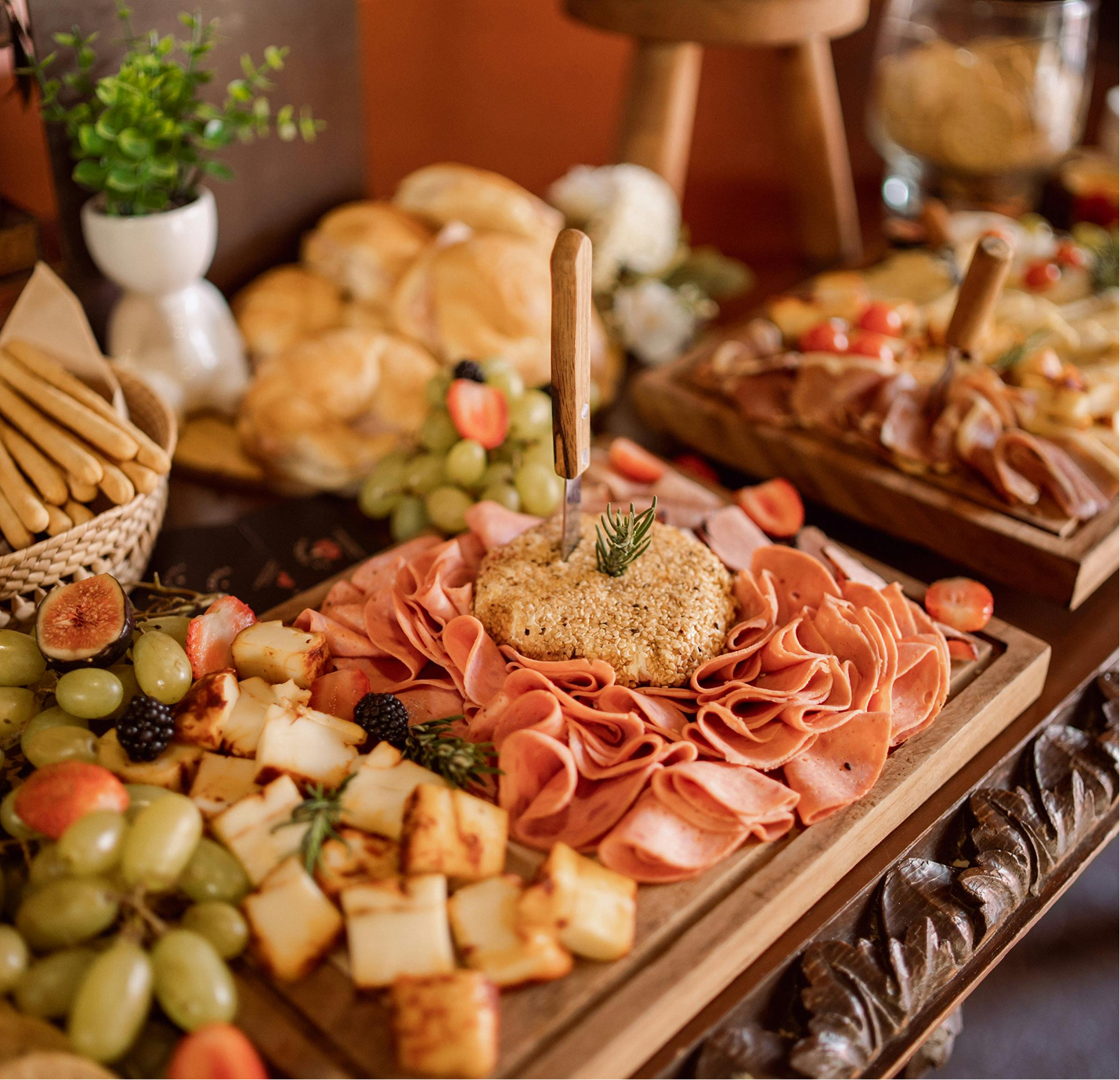 A cheese and meat platter with grapes, figs, and bread rolls, featuring a central round cheese with sliced meats arranged around it, garnished with a sprig of rosemary.