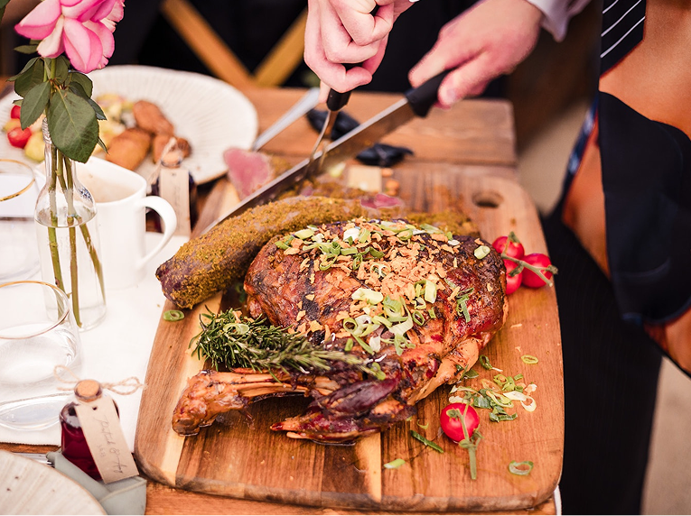 A roasted turkey with chopped herbs and vegetables on a wooden cutting board, next to fried breaded meat, with a chef carving. Surrounding items include a vase with pink flowers, glasses, a white mug, and cherry tomatoes.