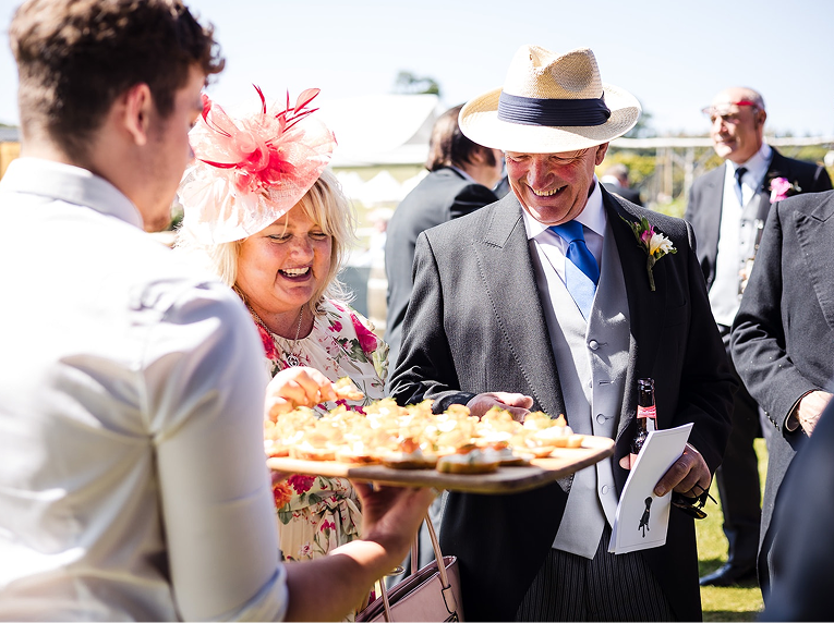 People celebrating at an outdoor wedding reception, with a woman in a floral dress and pink hat, and a man in a suit and straw hat, smiling as a server offers food.