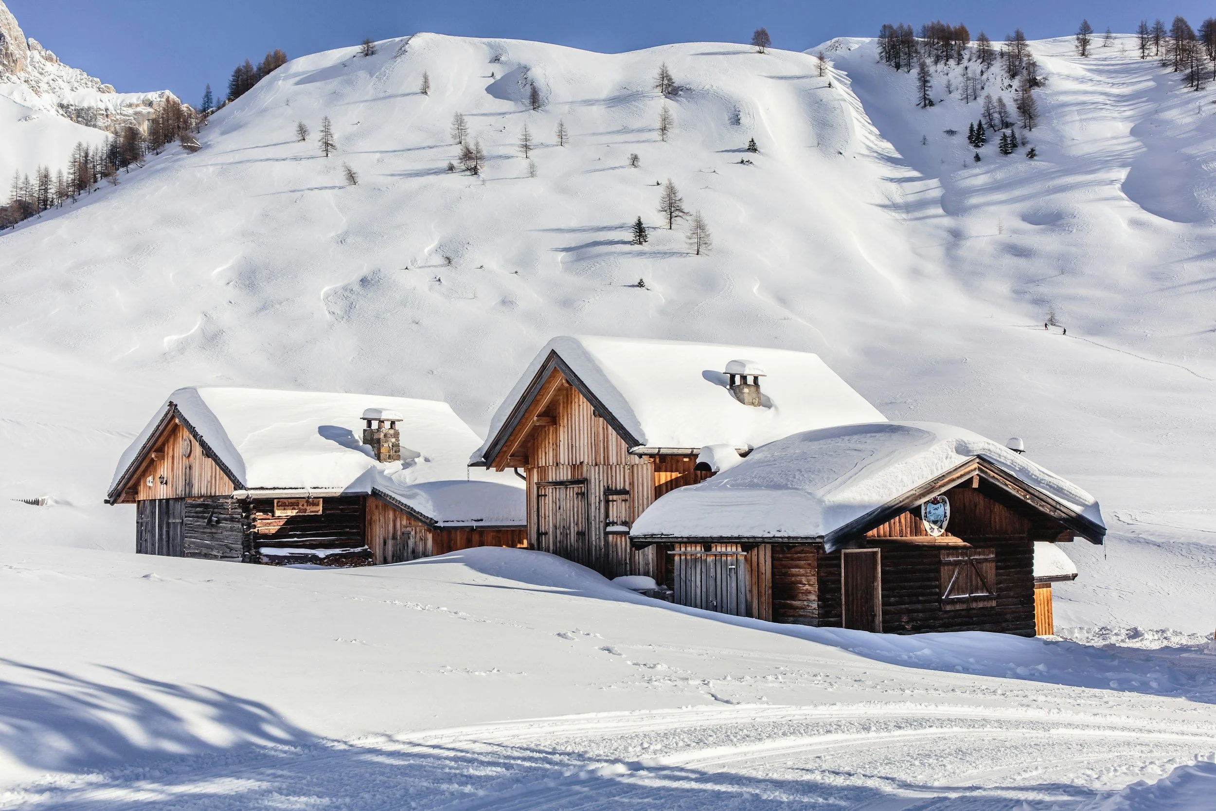 A wooden cabin surrounded by snow in a mountainous landscape with more snow-covered hills and trees in the background.