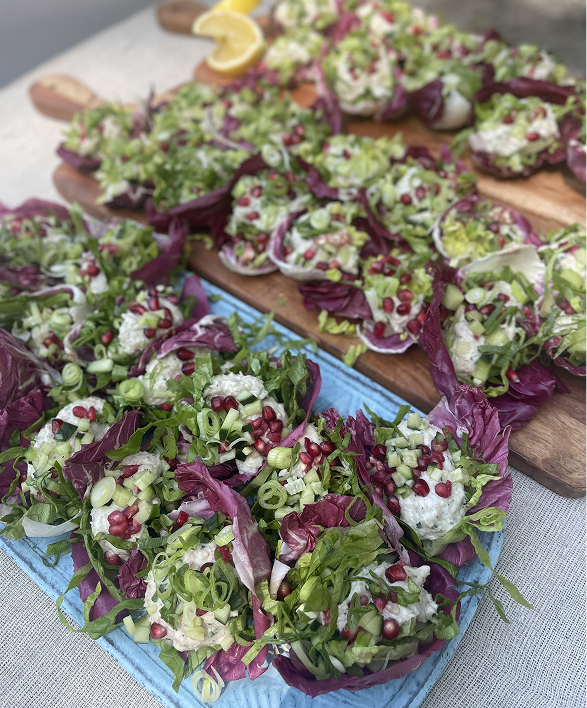 Tray of stuffed lettuce cups topped with chopped green onions, pomegranate seeds, and fresh herbs, garnished with purple lettuce leaves, served on a blue platter with a wooden cutting board in the background.