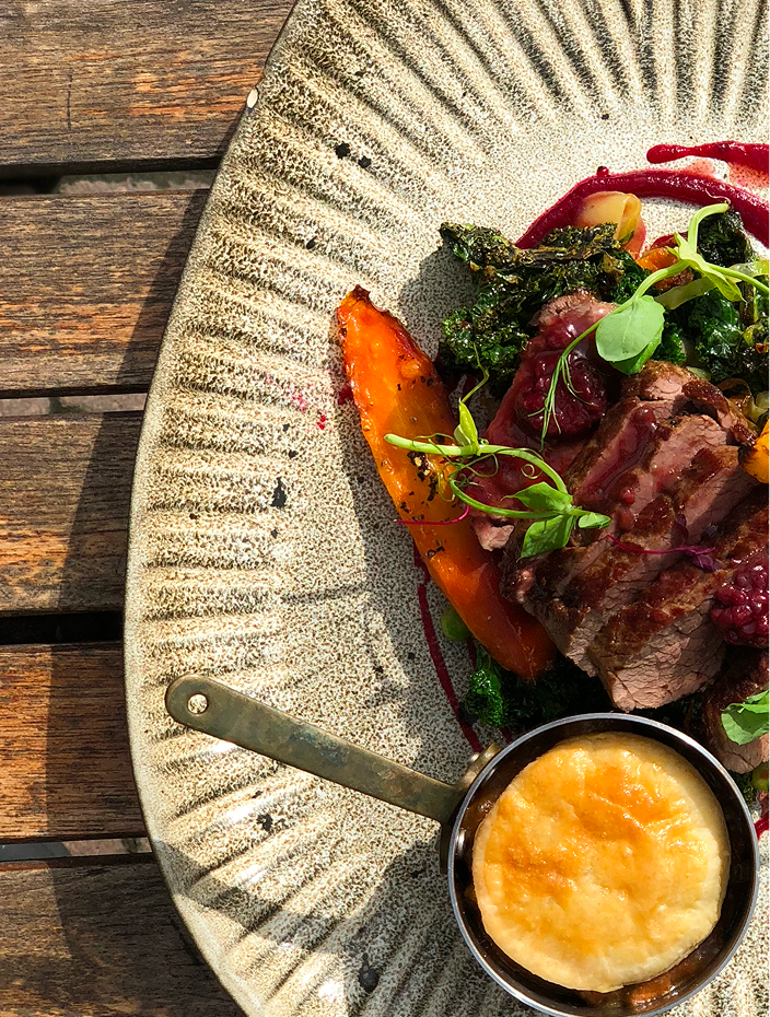 A plate of grilled meat with roasted vegetables, greens, and a small cup of mashed potatoes with gravy.