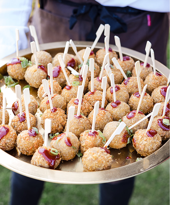 Tray of appetizers with breaded balls topped with sauce and green garnish, each on a stick.