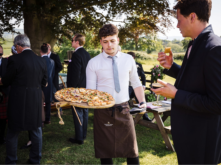 Young man in a white shirt and gray tie holding a tray of assorted appetizers at an outdoor event with other guests in formal attire.