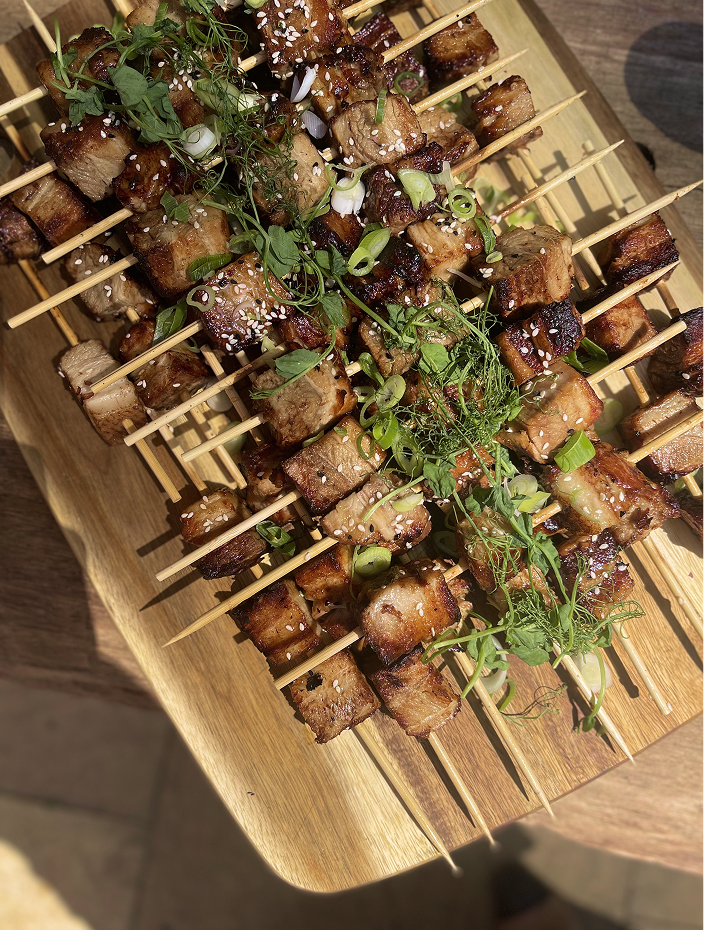 Close-up of grilled skewers with meat, garnished with green herbs, chopped green onions, and sesame seeds on a wooden platter.