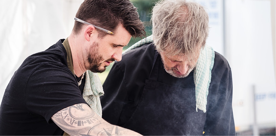 Two men cooking together, one appears younger with dark hair and tattoos, the other older with gray hair and a beard, both wearing black shirts and aprons, working in a kitchen.